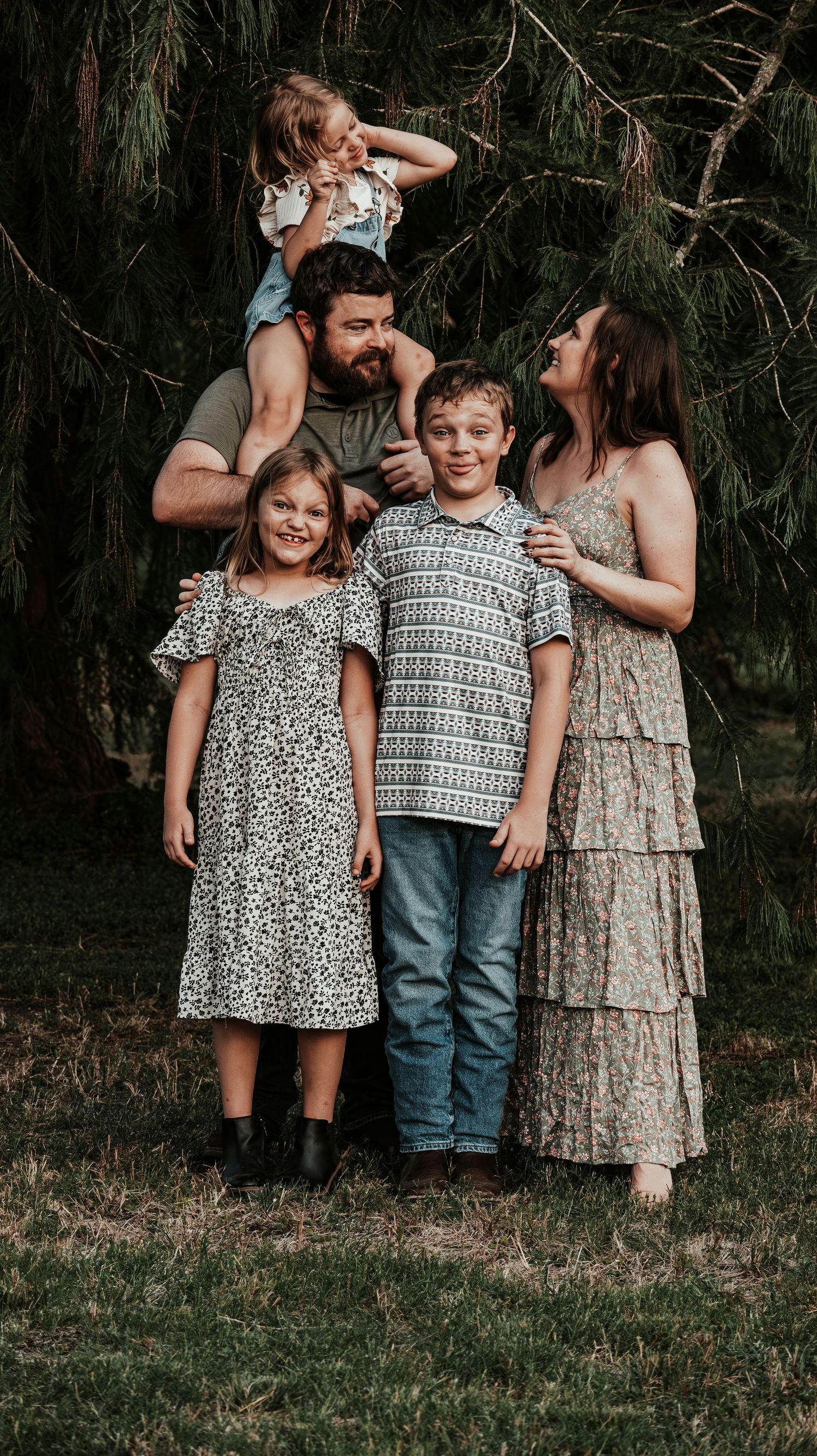 Joyful family of five poses outdoors with lush pine trees, toddler playfully perched on fathers shoulders.