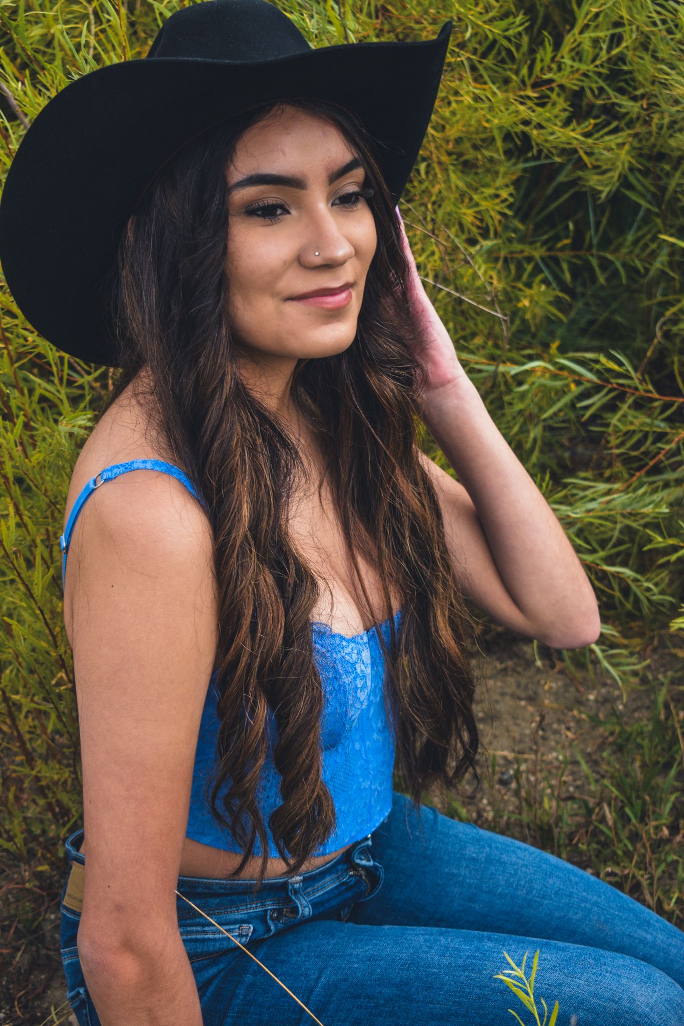 Beautiful young woman in black cowboy hat and blue lace crop top sits outdoors among green foliage for senior photos.