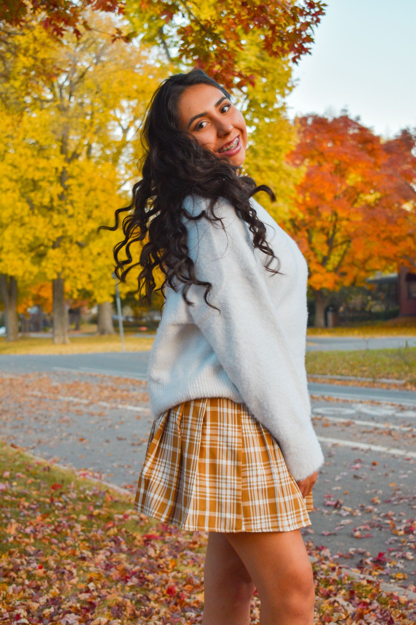 Smiling young woman in white sweater and plaid skirt twirling near colorful fall foliage in park for senior photos.