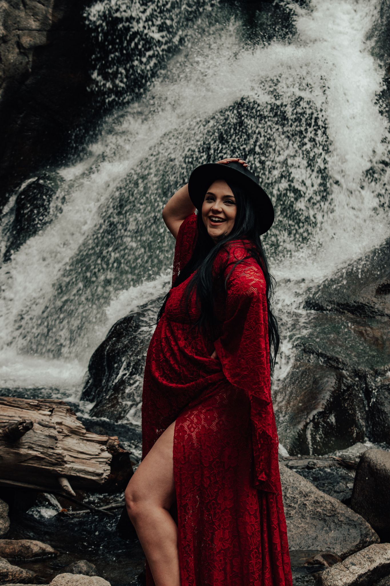 Pregnant woman in red lace gown and black hat poses on rocks in front of a rushing waterfall. Maternity Photographer in Pflugerville, Tx