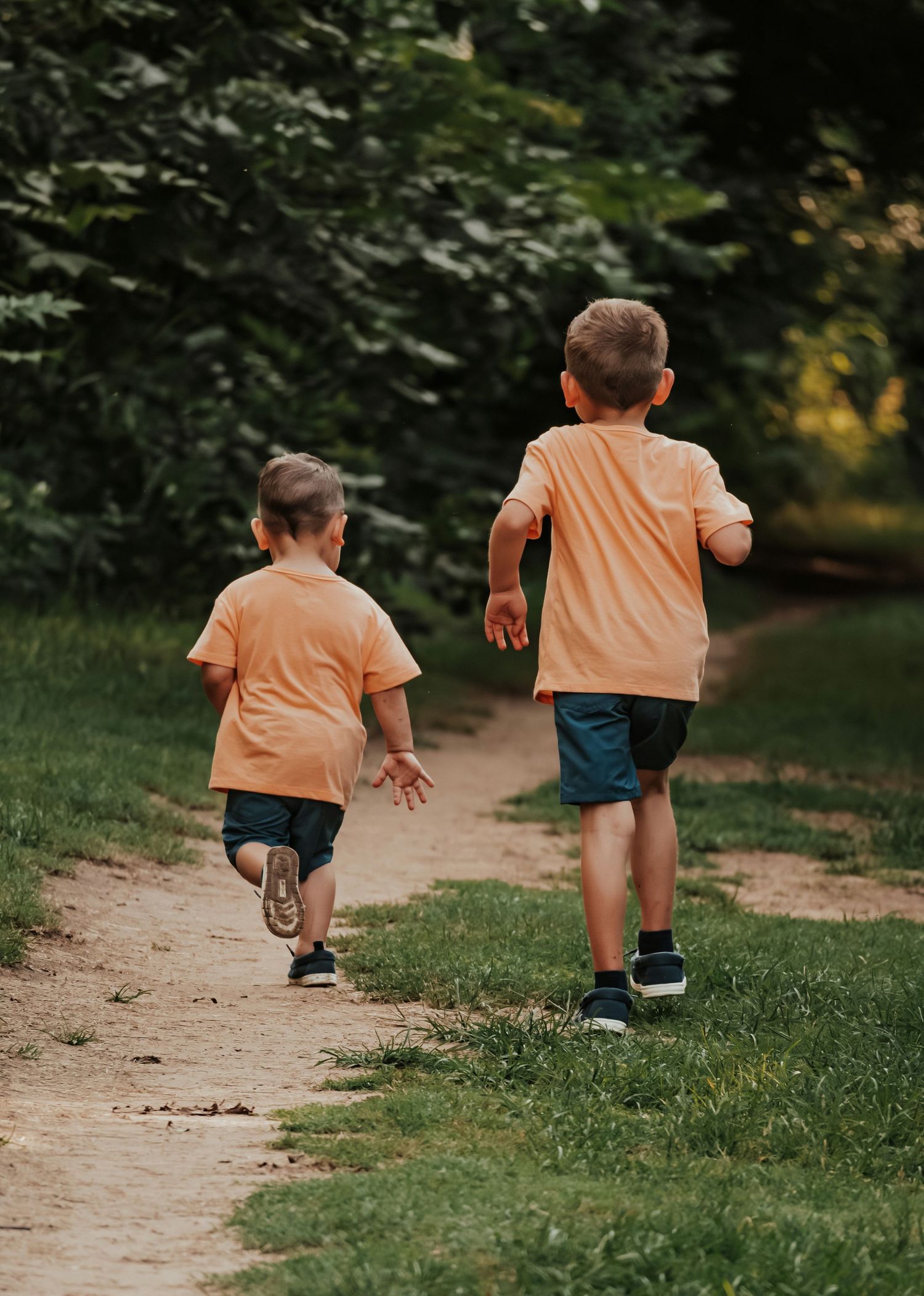 Two young brothers in matching orange shirts and blue shorts run together along a park path.