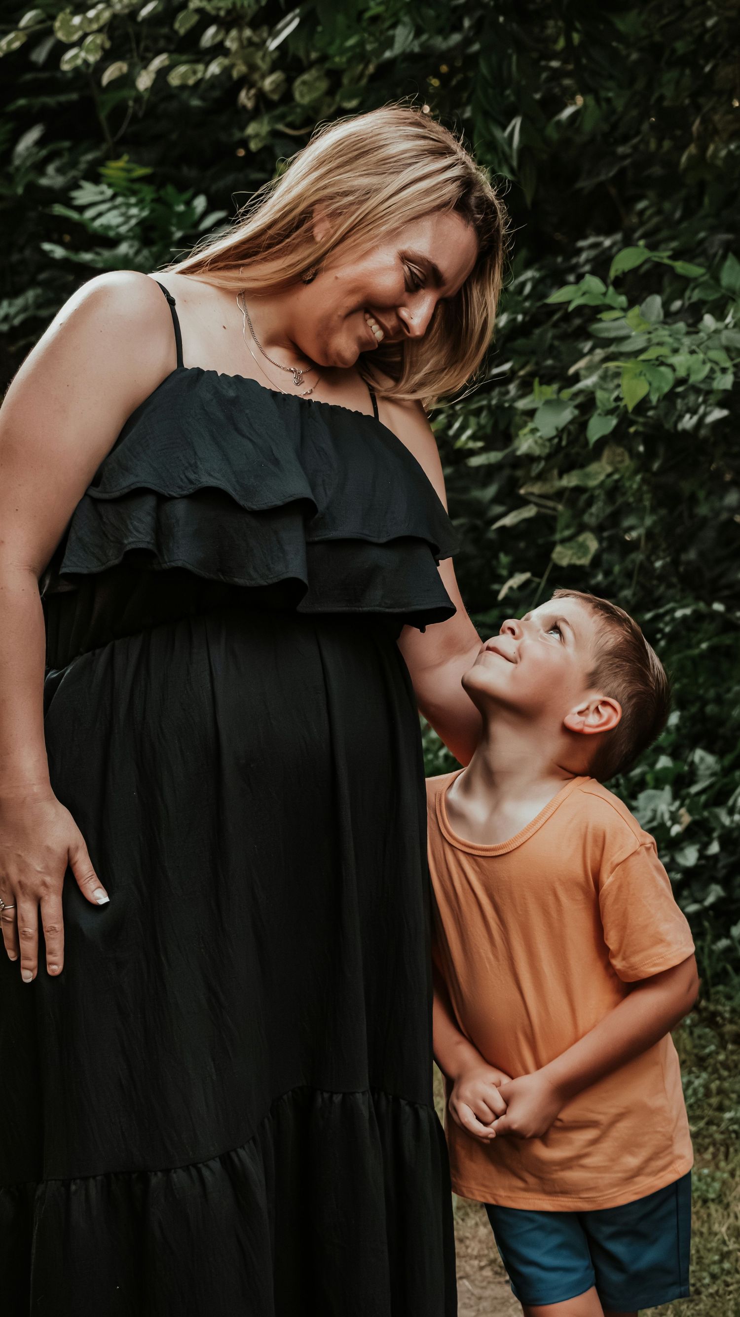 Pregnant woman in black dress smiling down at young boy in orange shirt outdoors.
