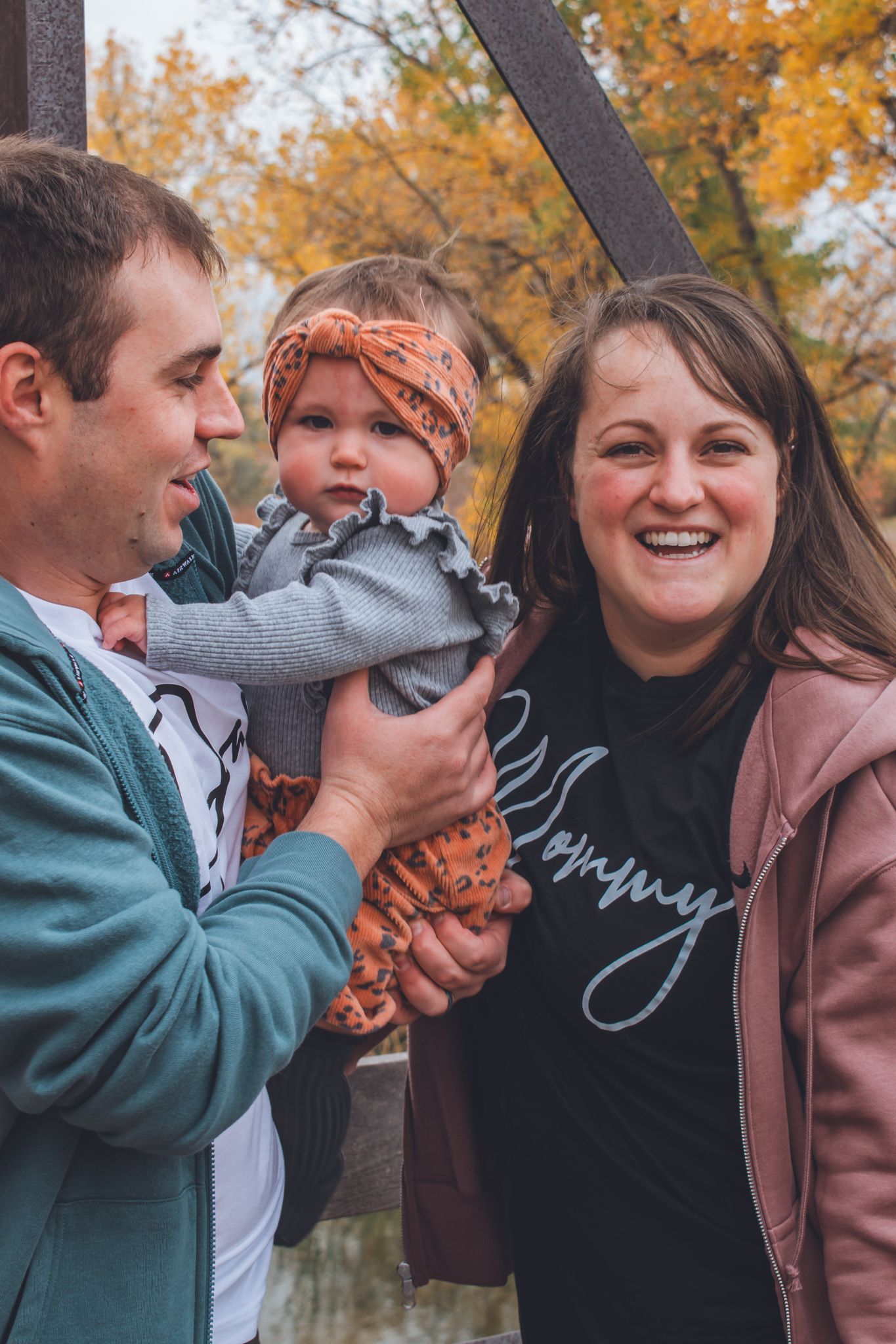 Happy family with a smiling mother wearing a Mommy shirt holding their baby girl in autumn park. Family Photographer in Pflugerville, Tx