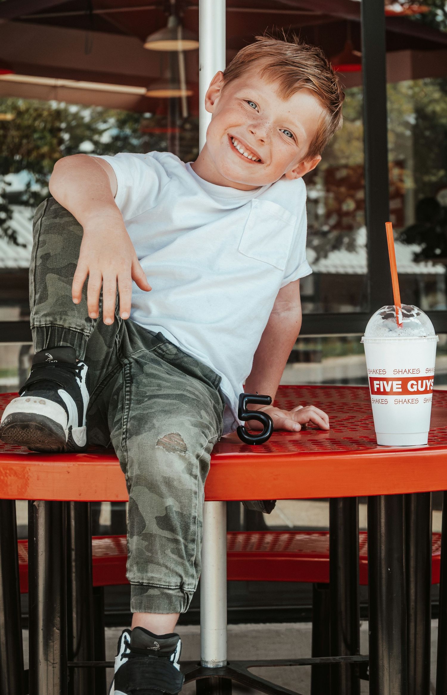 Smiling red-haired boy in camo pants sits at a Five Guys restaurant table with a milkshake drink. Family Photographer in Pflugerville, Tx