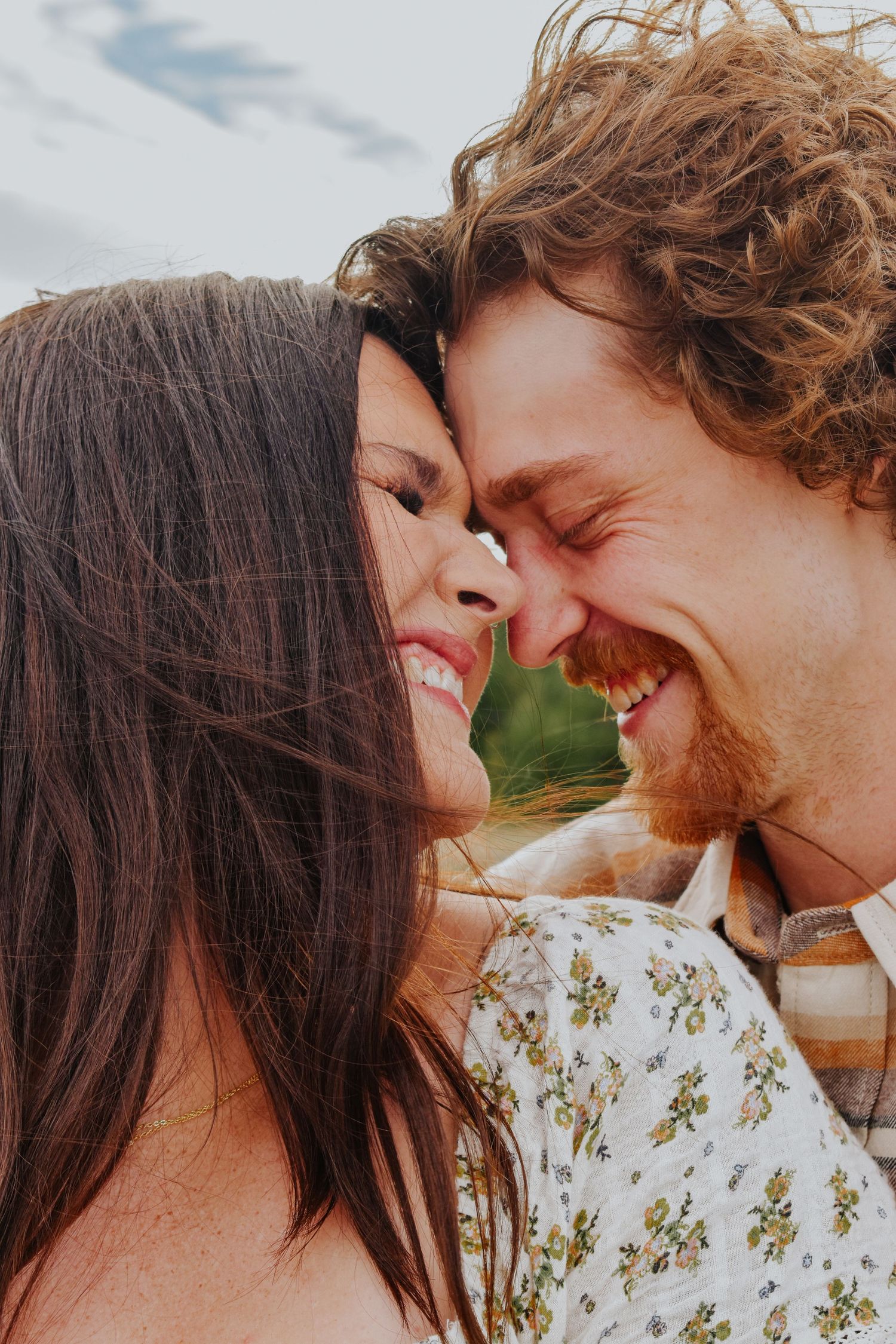 A joyful couple presses their foreheads together, both laughing in a bright outdoor setting.