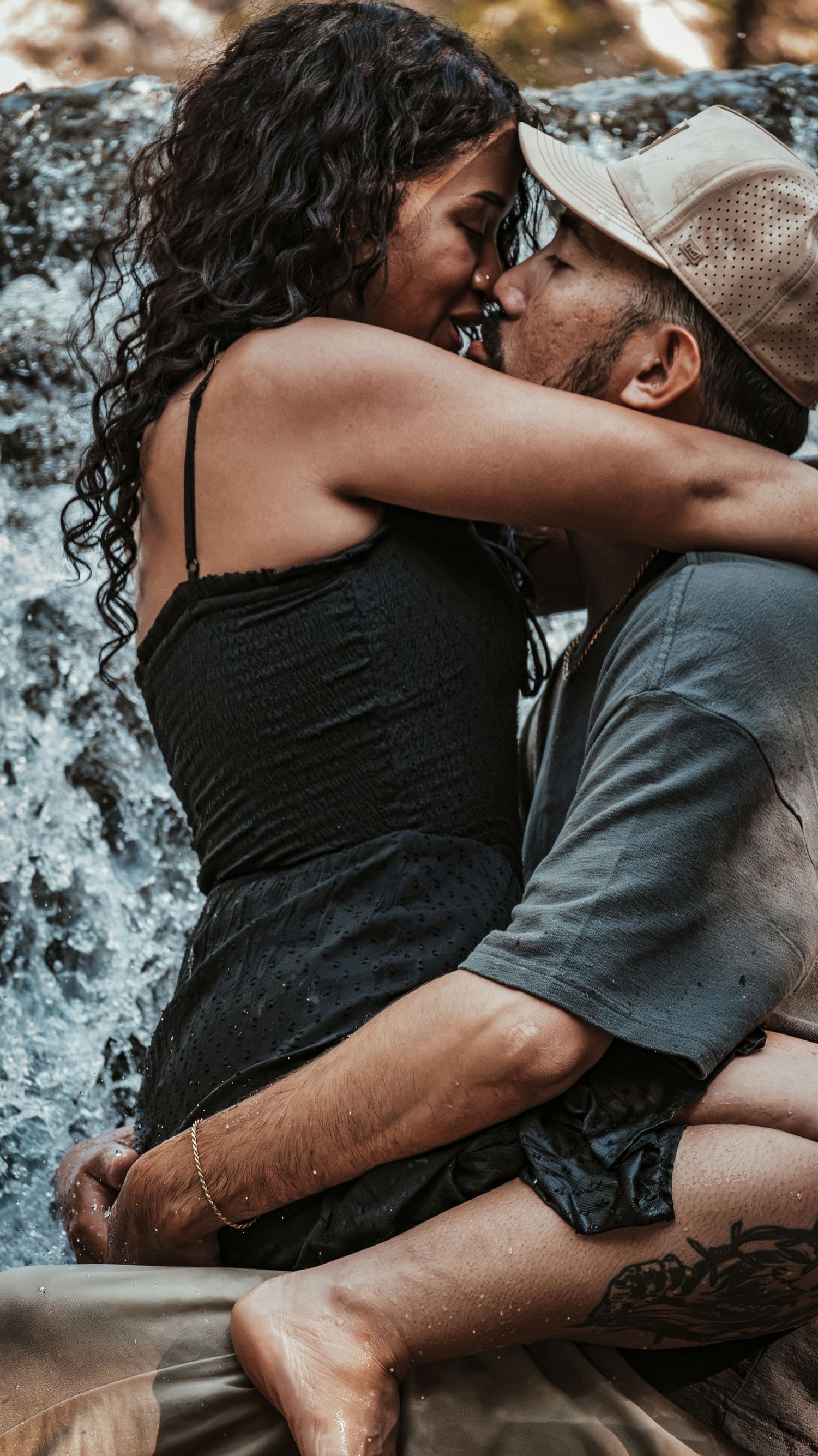 Romantic couple kissing intimately while seated on rocks beside a flowing waterfall.
