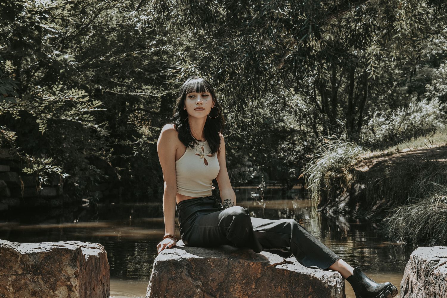 Young woman with dark bangs sits on a large rock by a peaceful forest creek on a sunny day for senior photos.