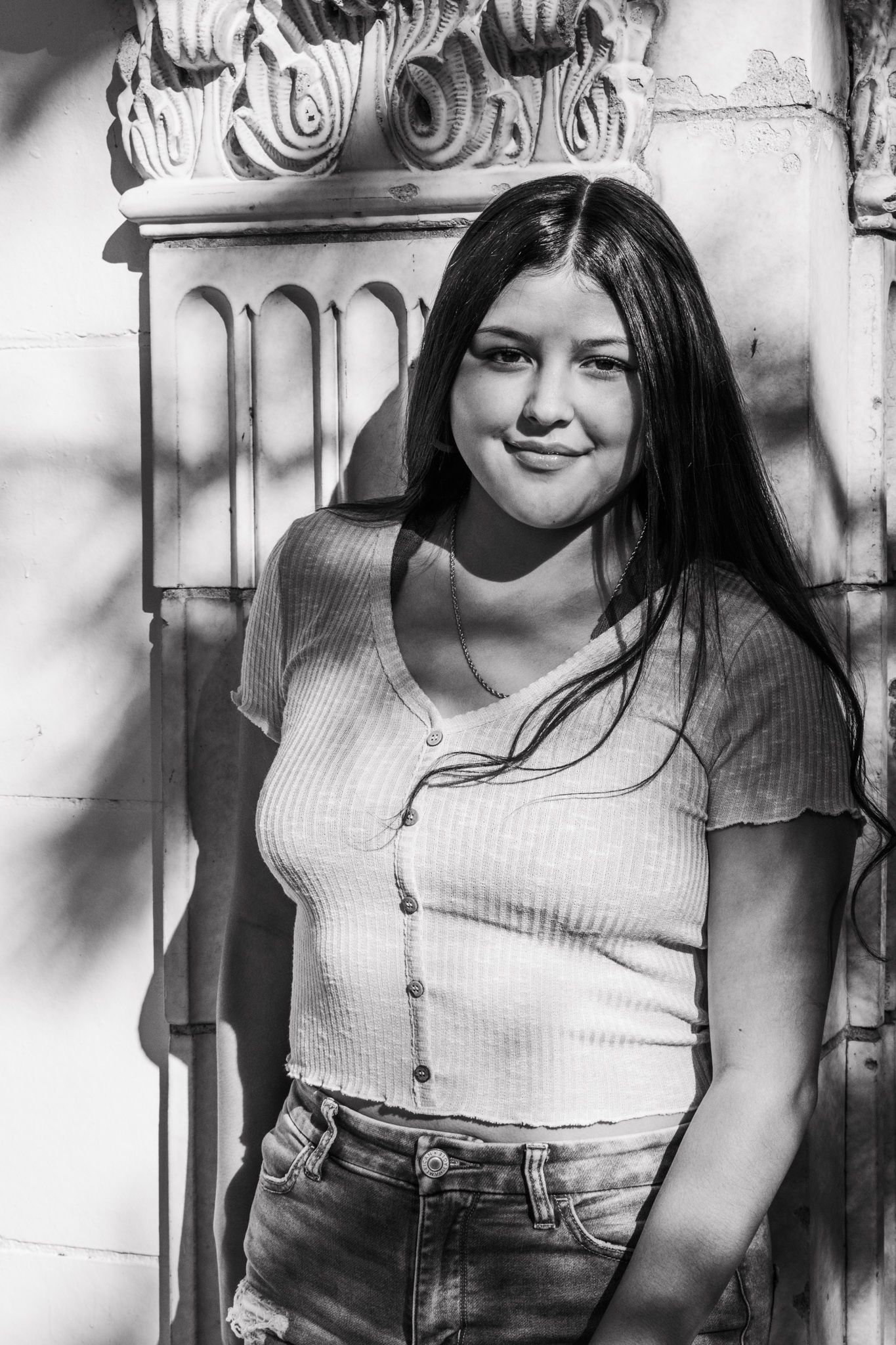 Black and white portrait of young woman leaning against decorative stone column for senior photos.