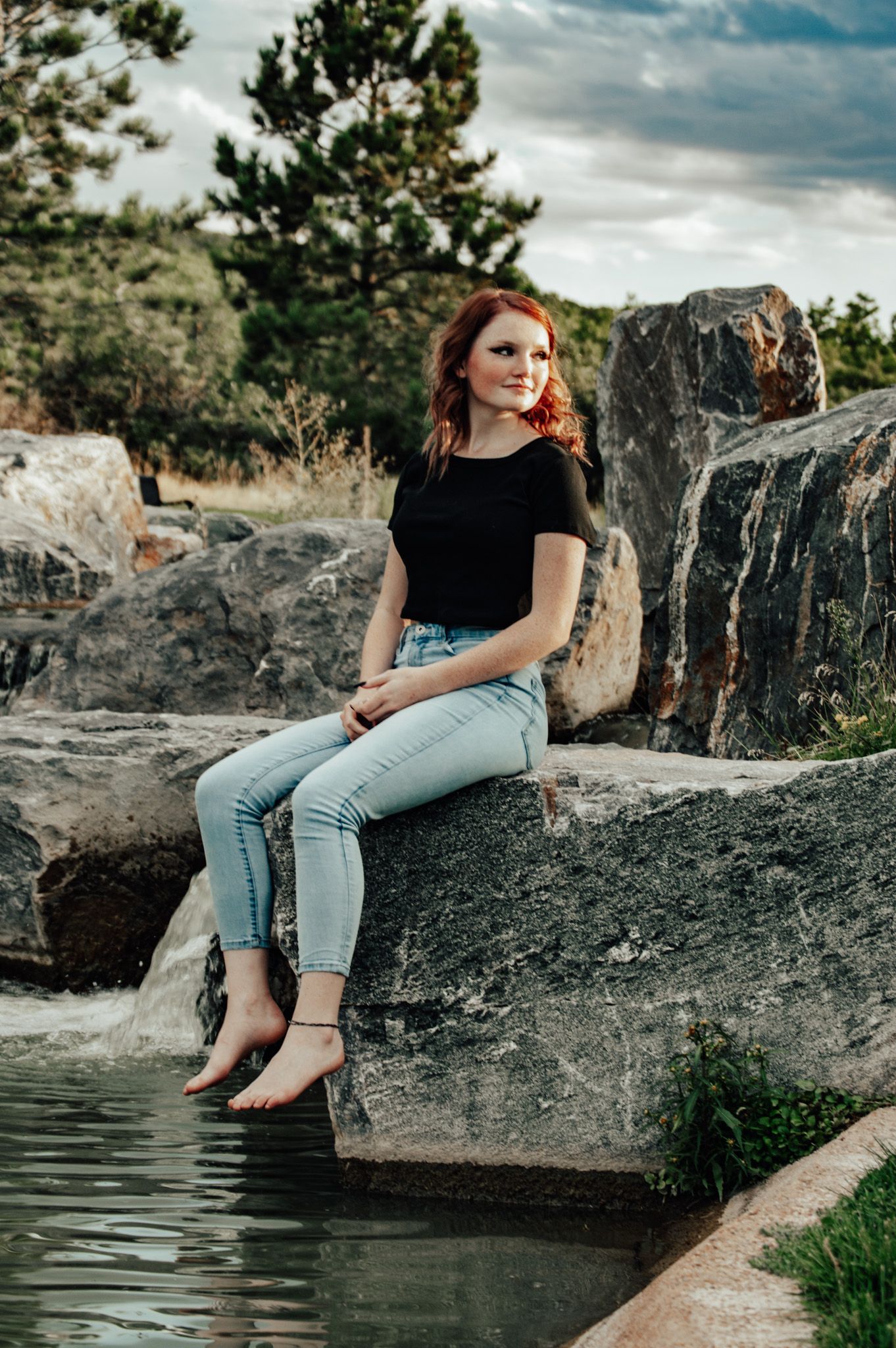 Redhead woman in black tee and light jeans sitting on large rock with bare feet near water outdoors for senior photos.