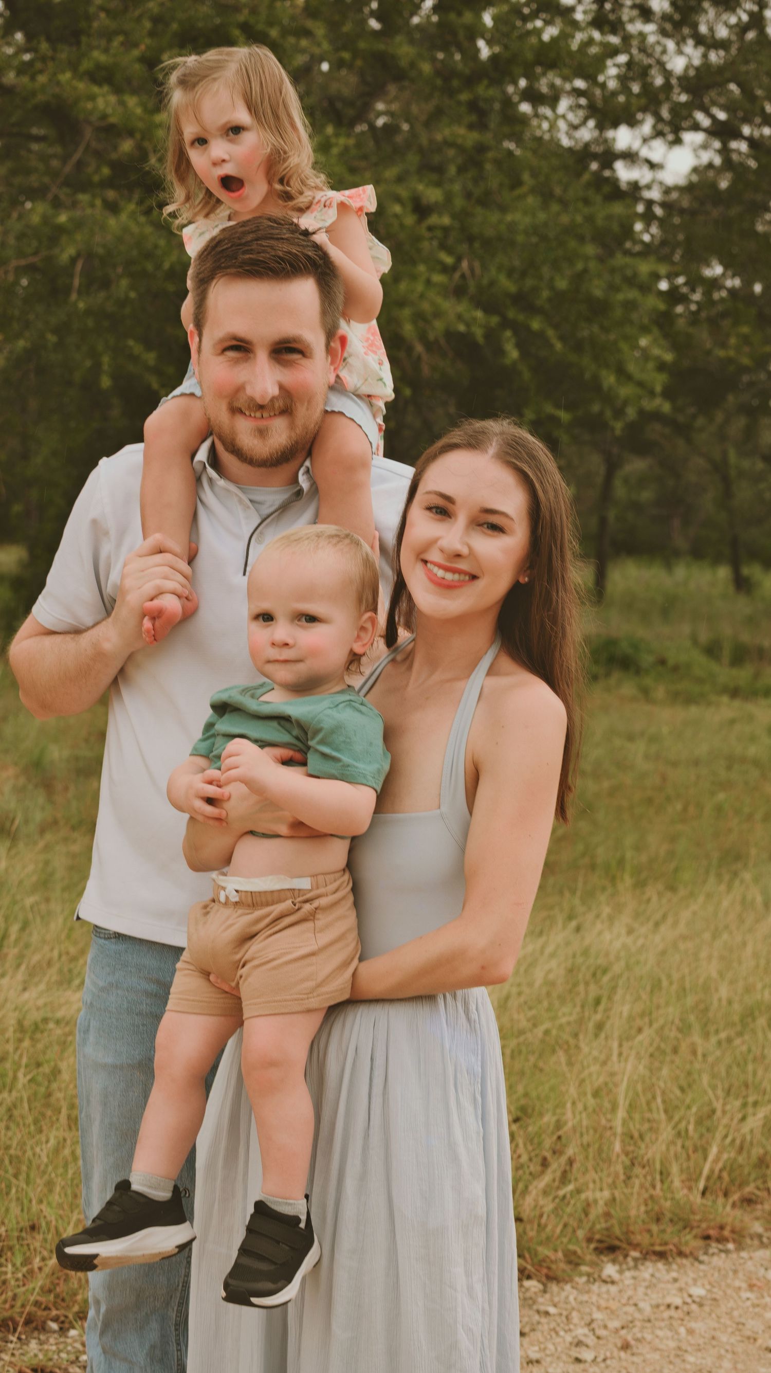 Happy family of four poses outdoors, parents holding two young children in a field.
