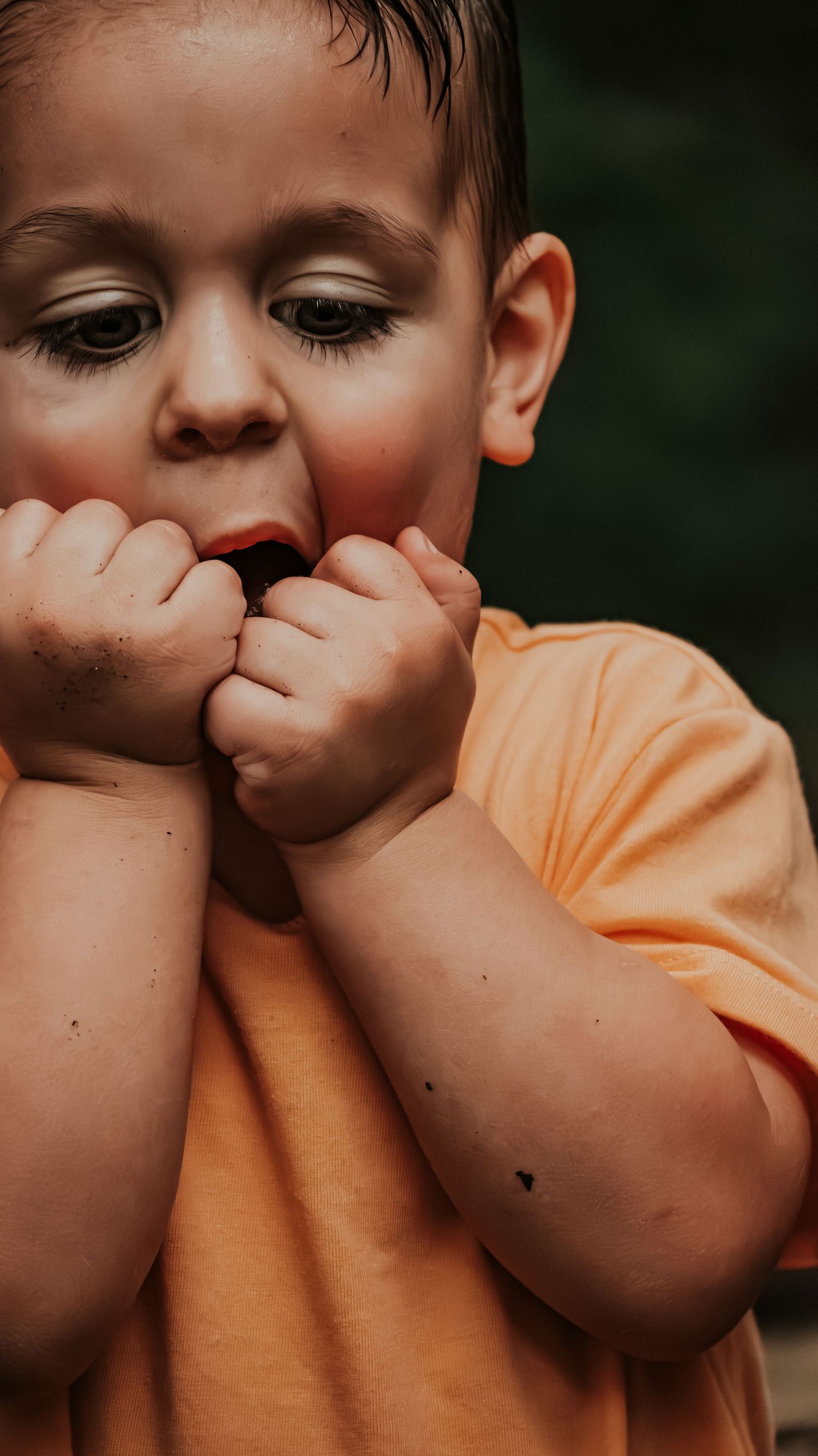 Young toddler boy in orange shirt biting his fists with a curious downward gaze outdoors.