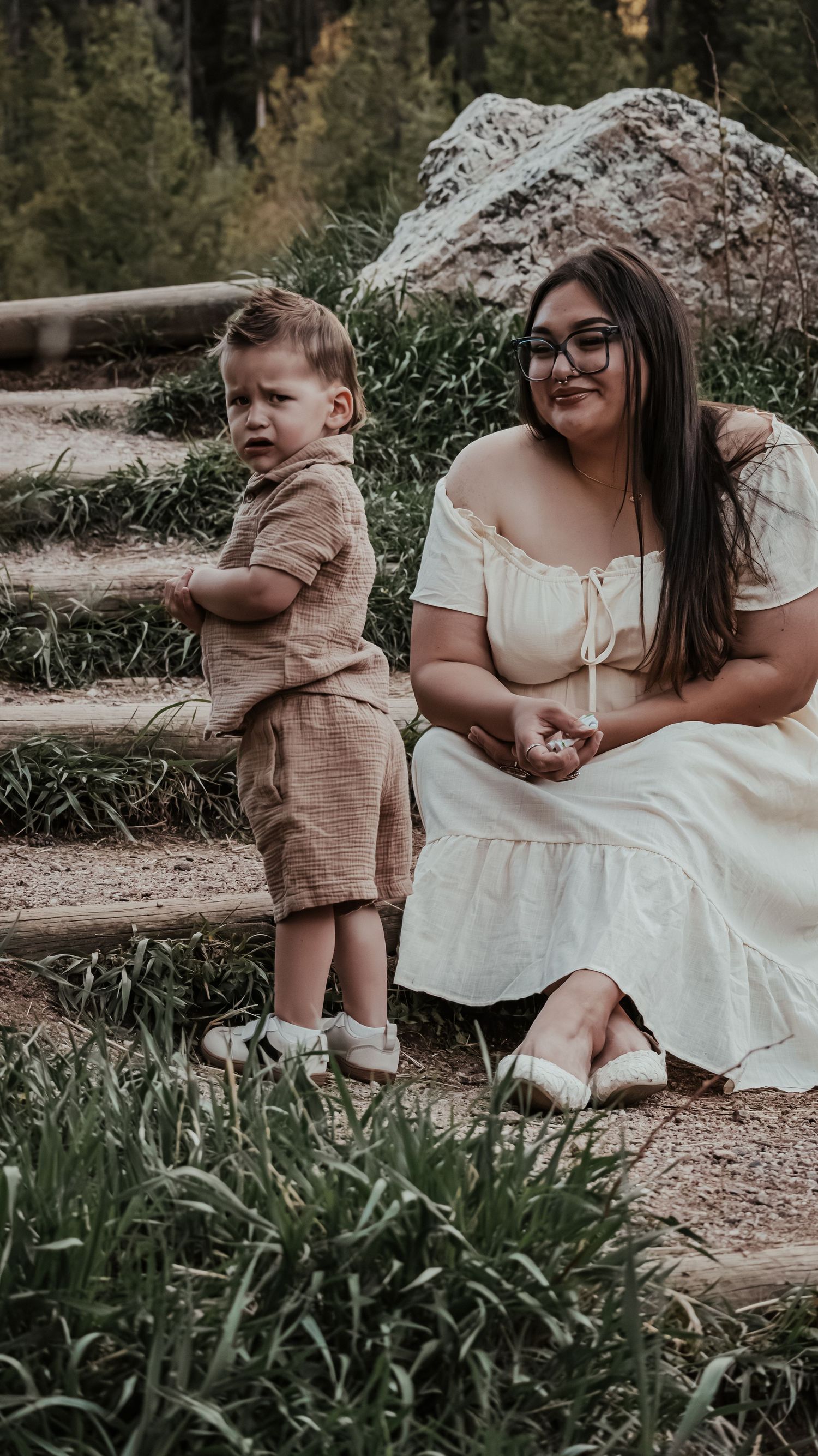 Young boy in tan outfit standing beside smiling woman in white off-shoulder dress on outdoor steps.