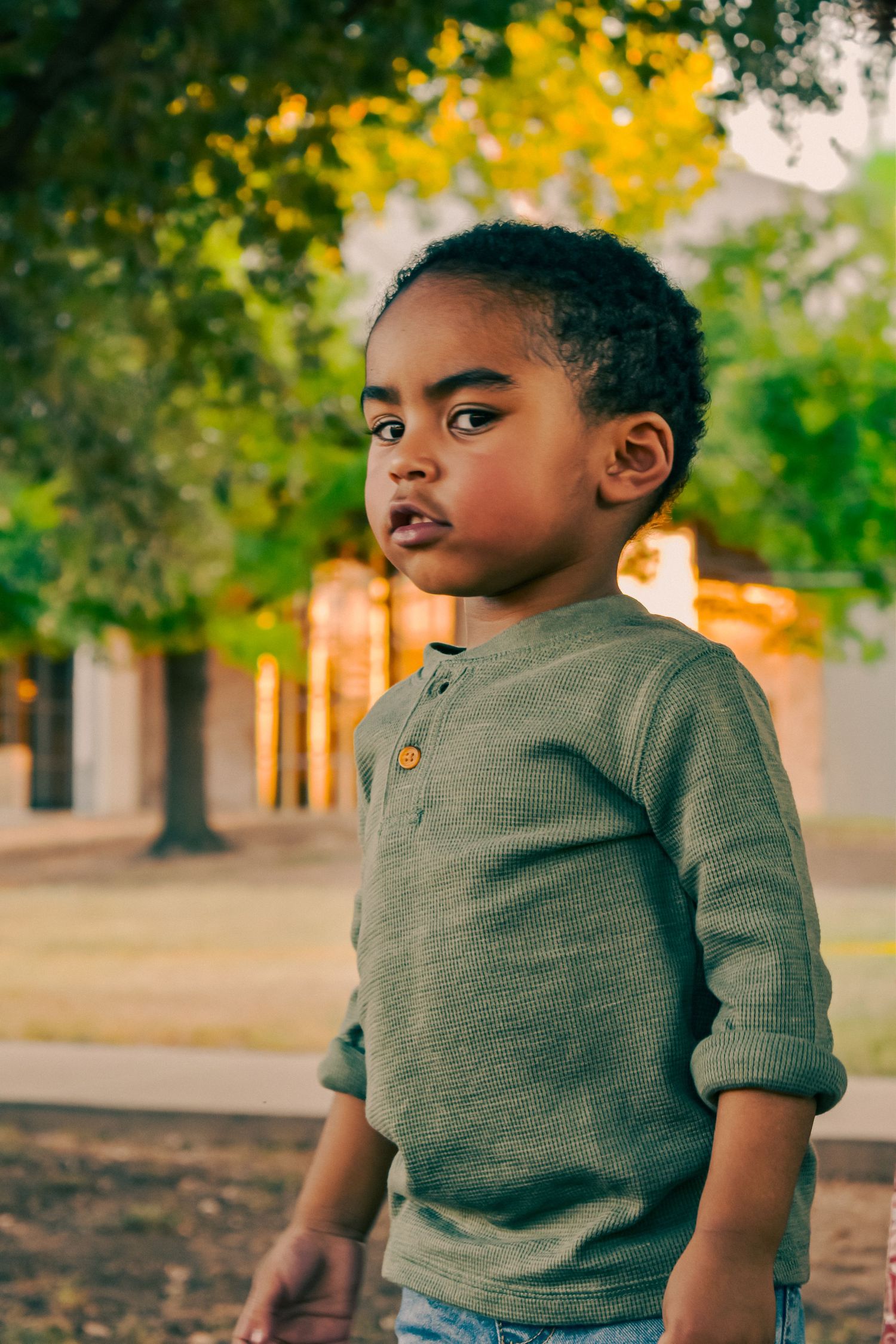Young Black toddler boy in green henley sweater standing outdoors with trees in background.