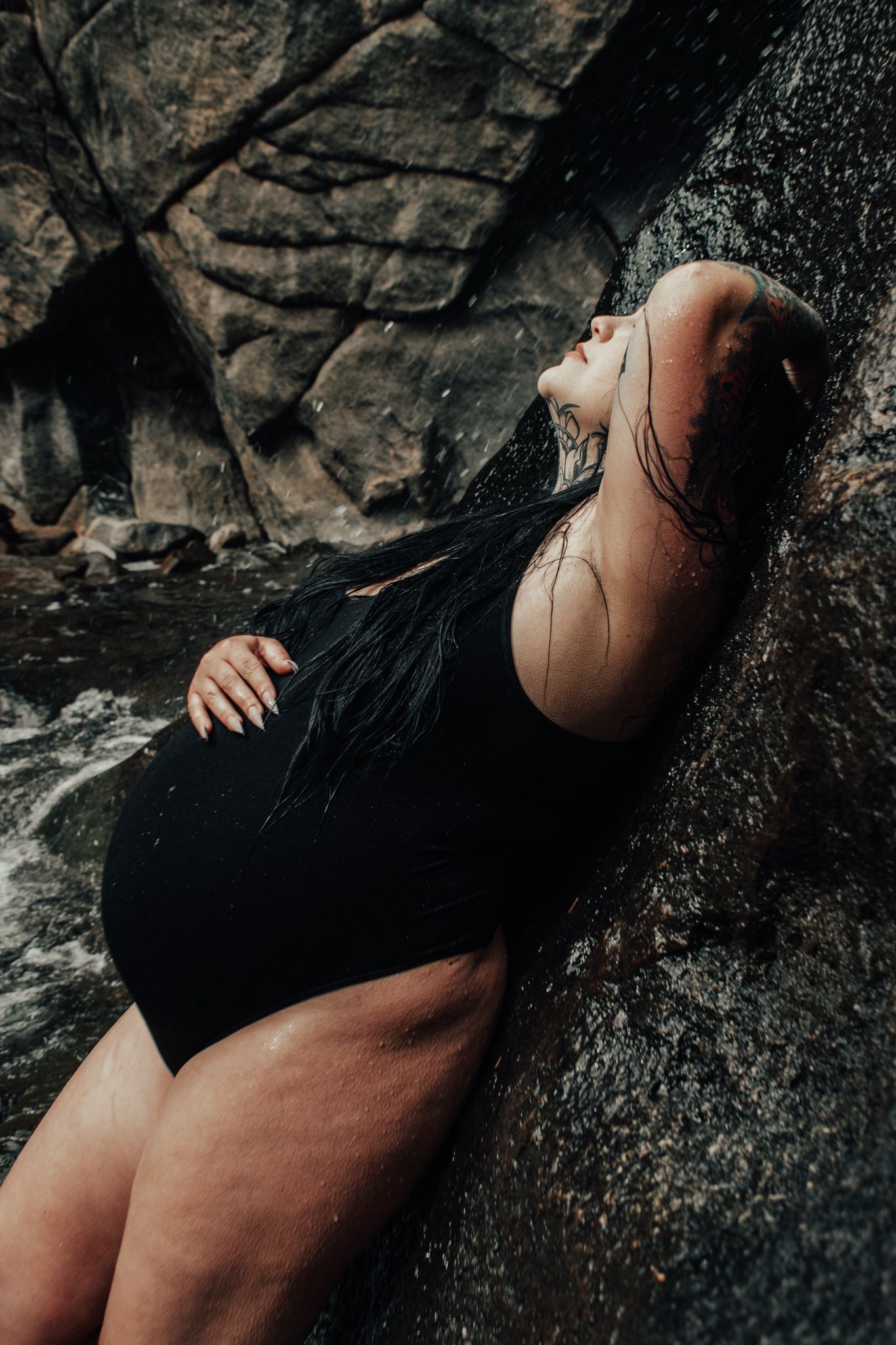 Pregnant woman with wet hair in black swimsuit leaning against mossy rock near waterfall.