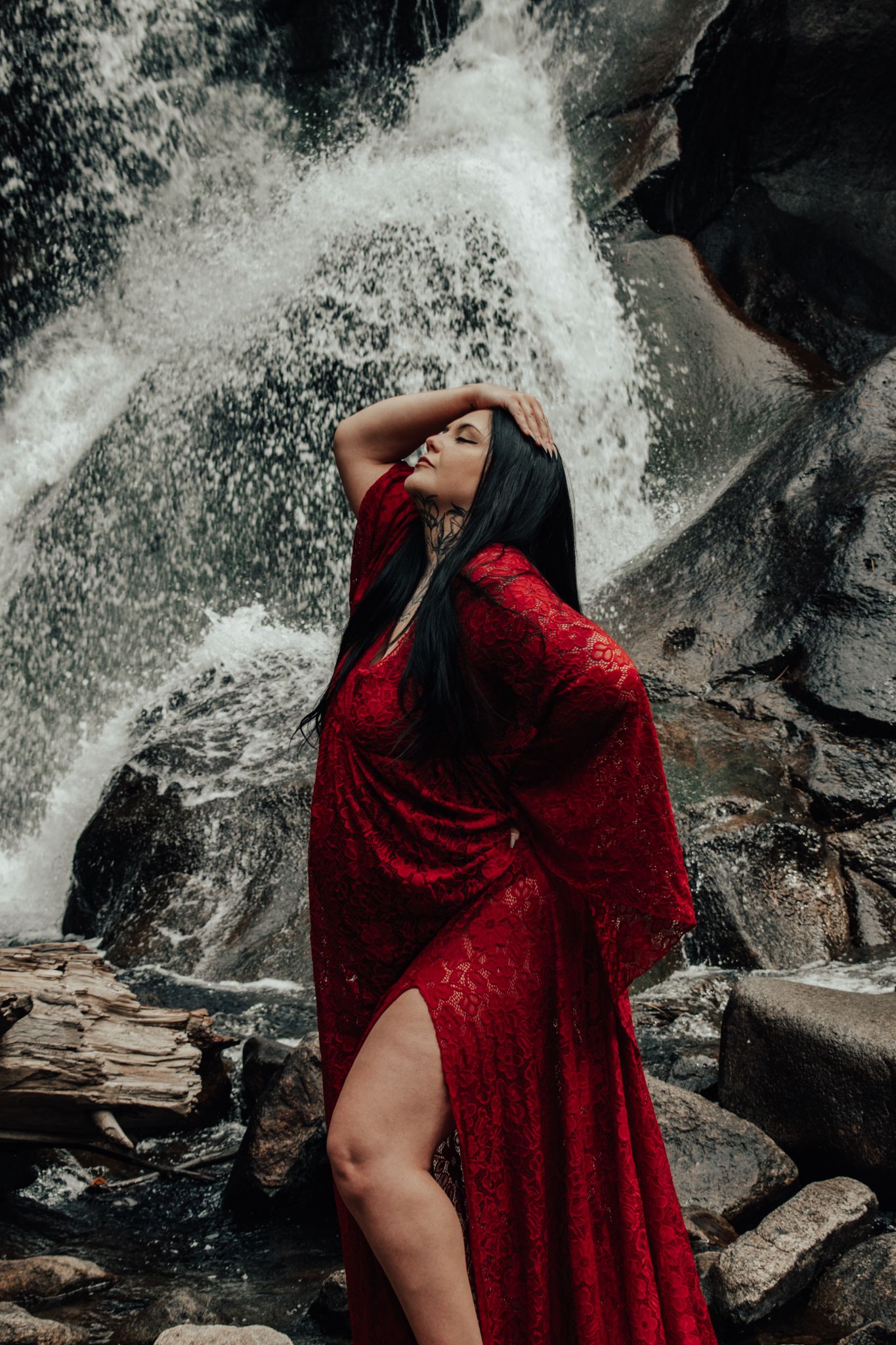Woman in elegant red gown with slit posing gracefully before a powerful cascading waterfall.