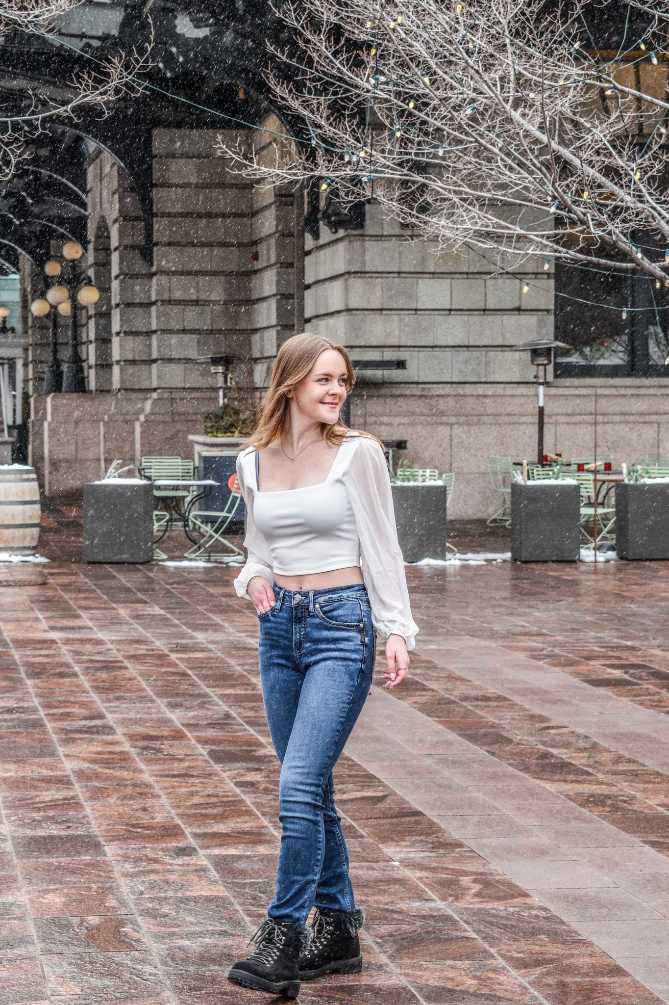 Young blonde woman in white crop top and jeans walking on wet brick plaza during light snowfall.