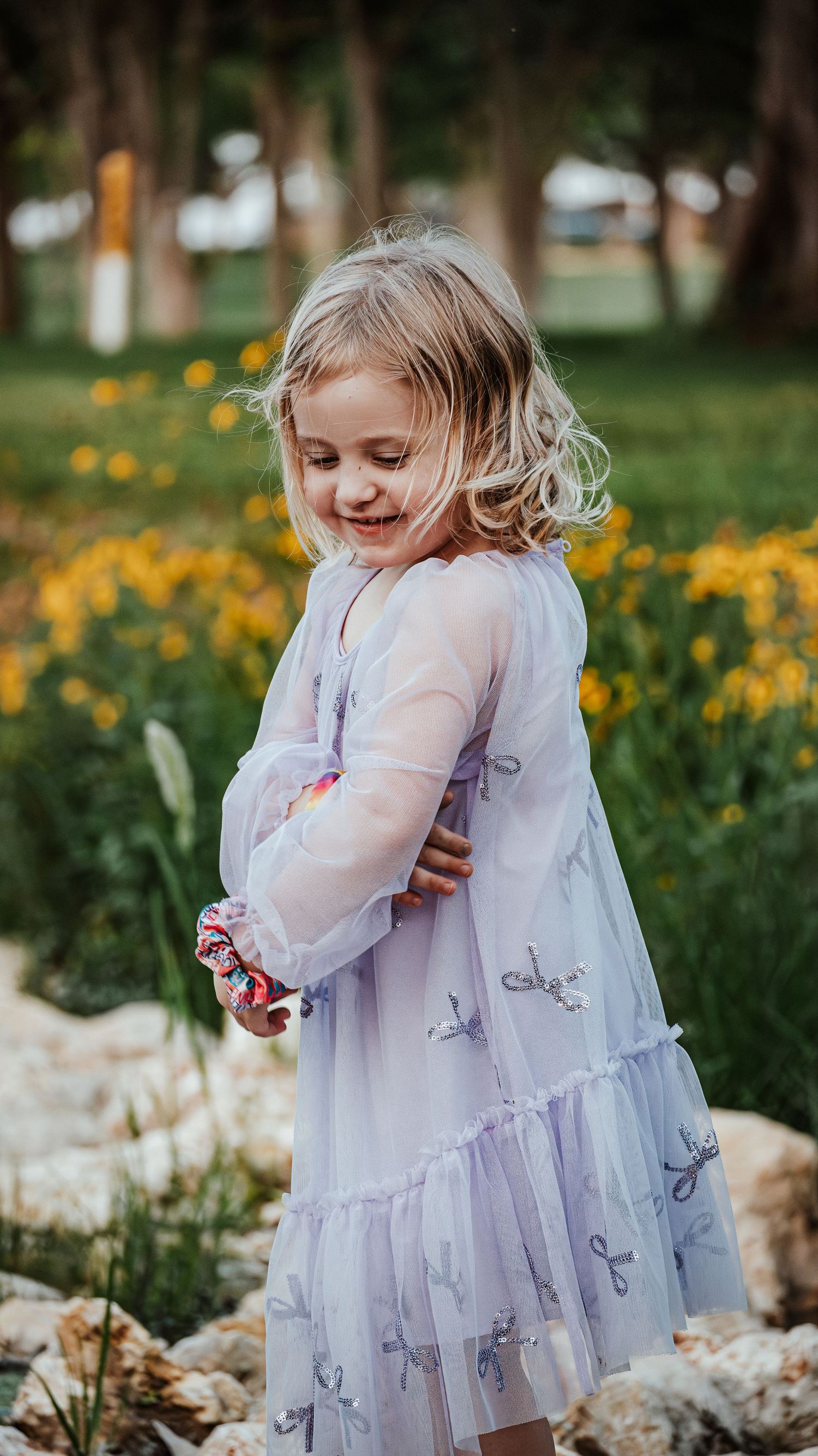 A smiling young girl in a light purple dress stands among yellow wildflowers in a garden setting.