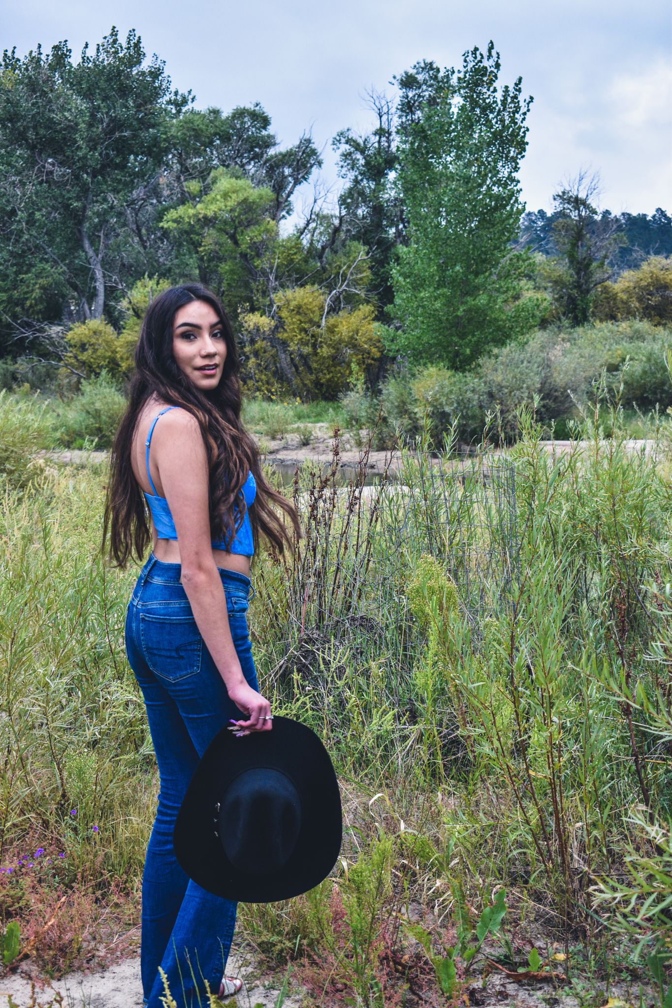 Young woman in blue crop top and jeans holds black cowboy hat in a lush green outdoor landscape for senior photos.