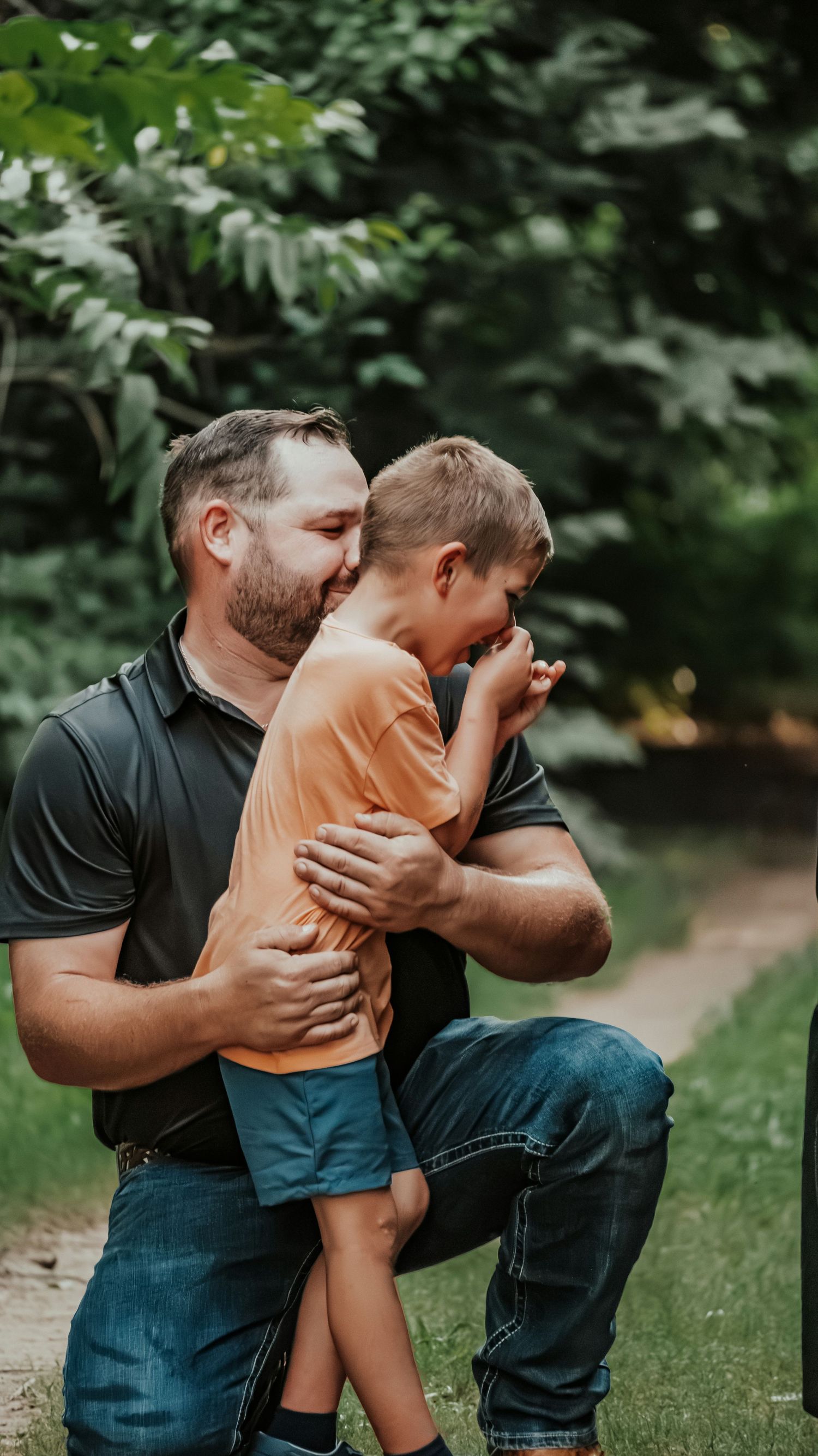 A man crouching down hugging a laughing young boy outdoors in a lush green park setting.