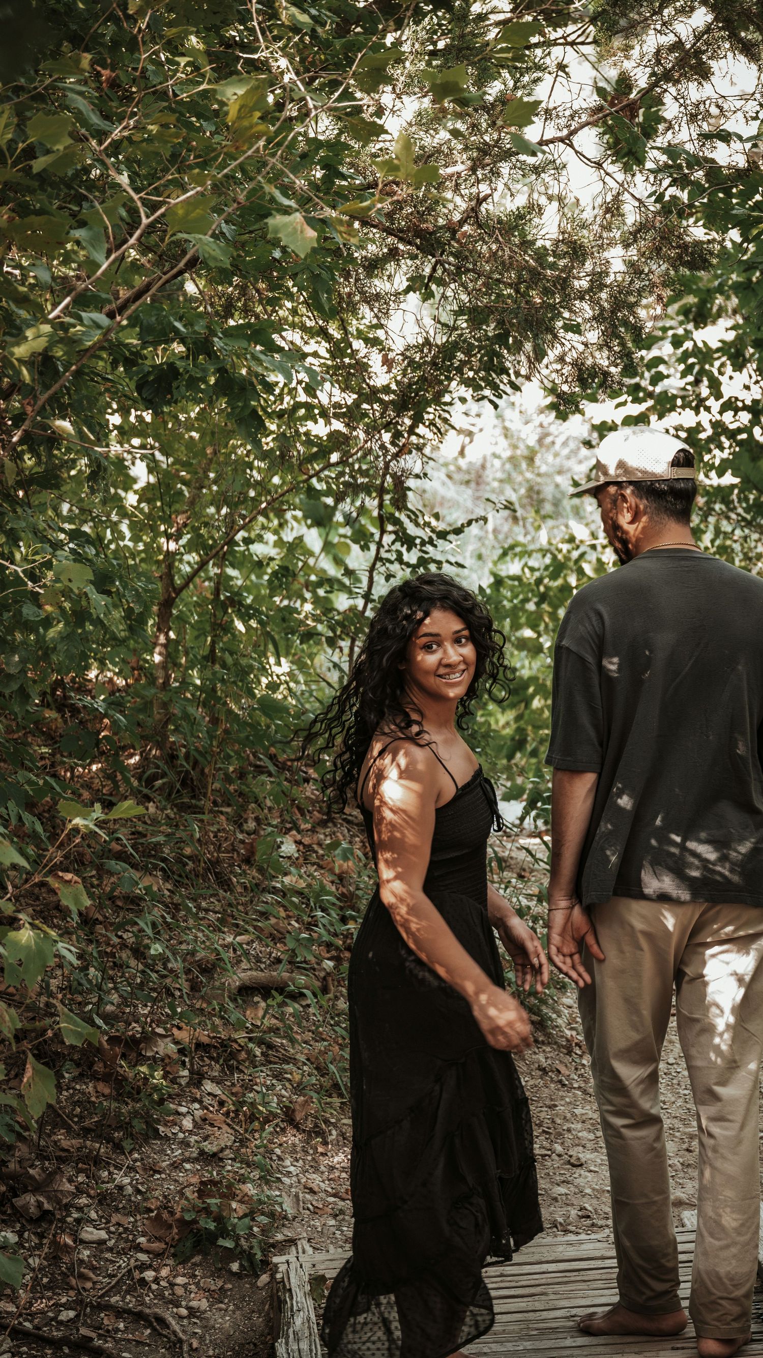 A couple holds hands on a wooden path surrounded by lush green trees for a couples session.