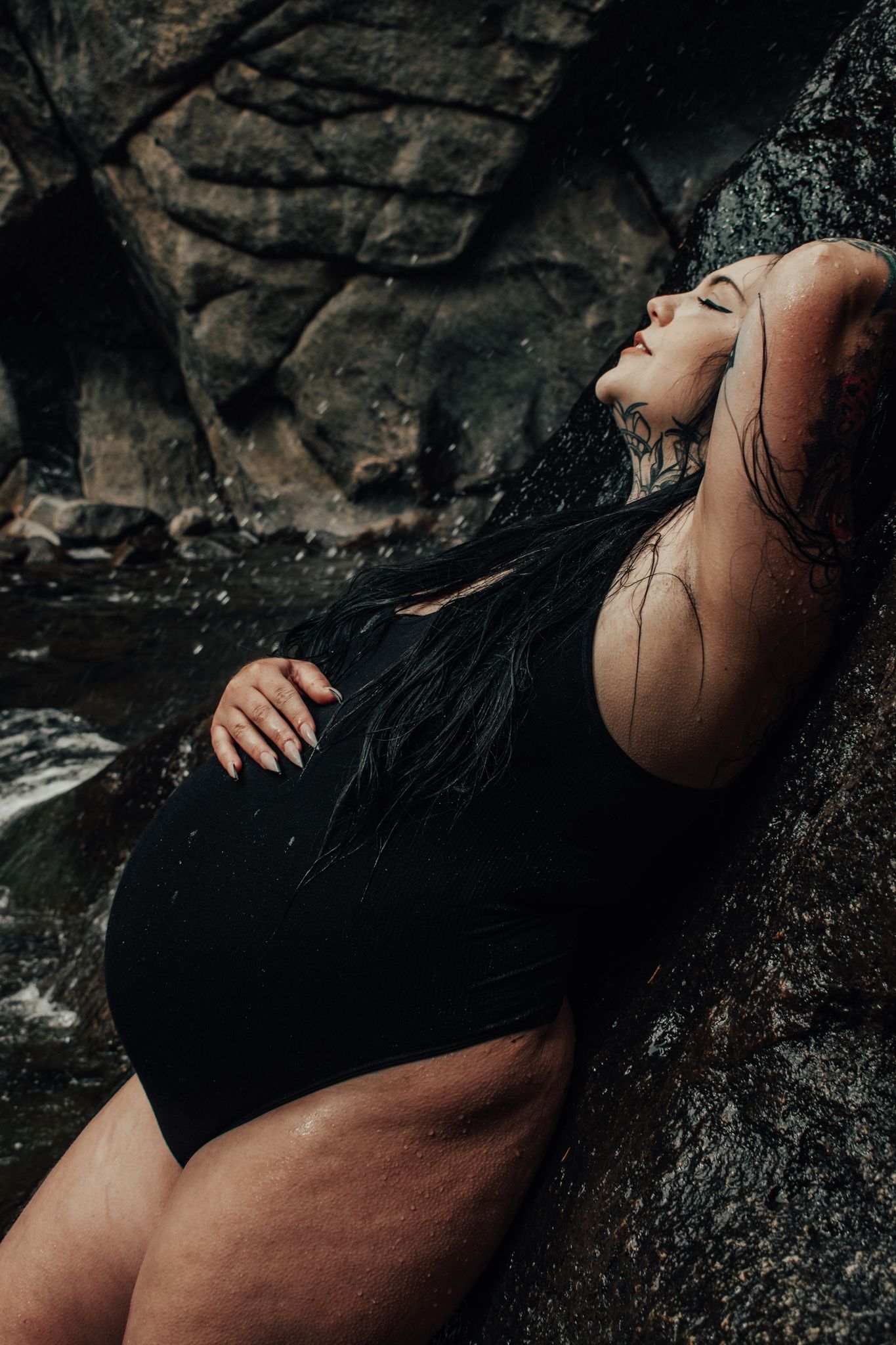 Tattooed pregnant woman in black swimsuit reclines against rocky surface near flowing water. Maternity Photographer in Pflugerville, Tx