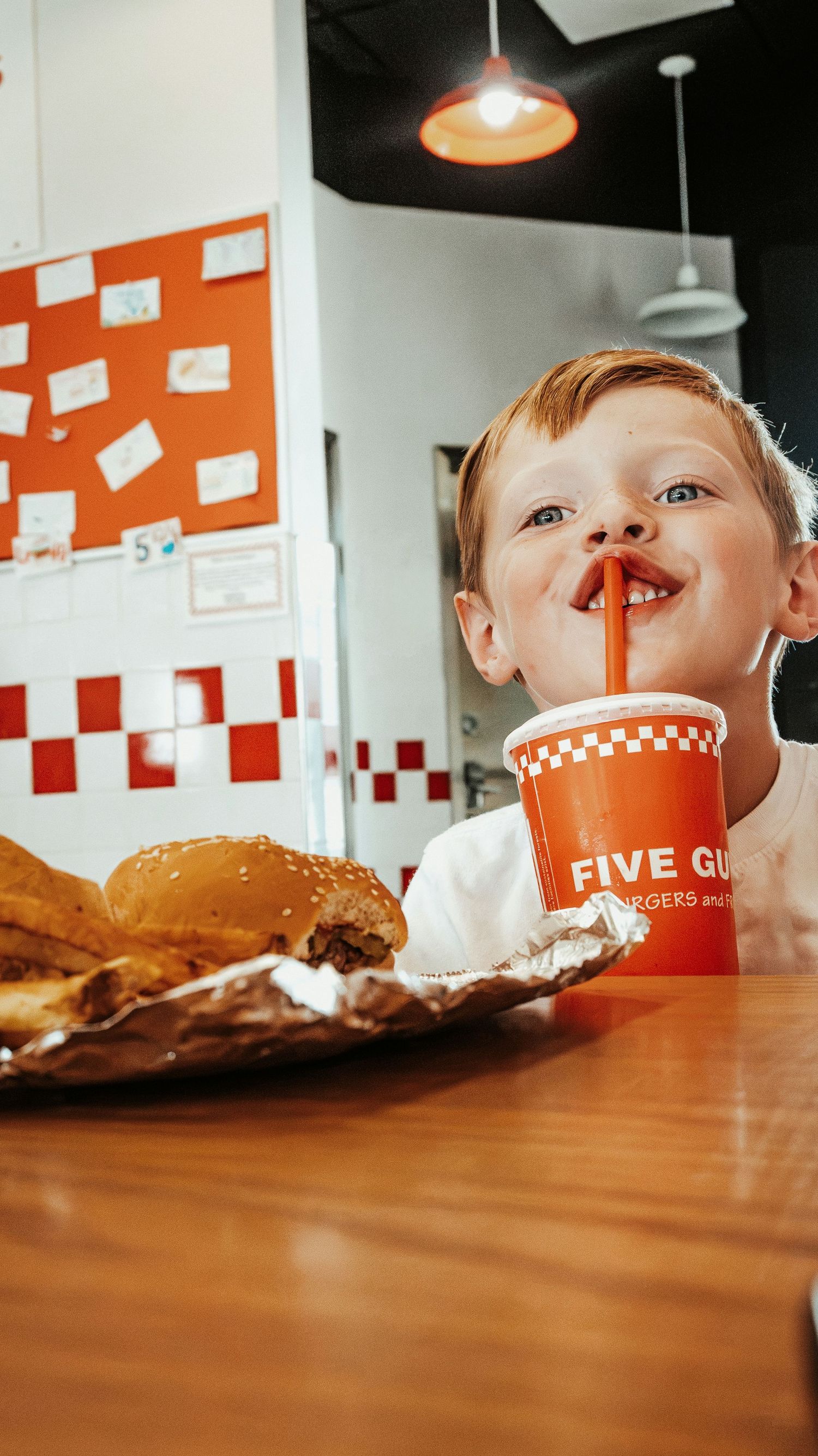 Happy young boy drinking soda through straw at Five Guys burger restaurant with meal on table. Family Photographer in Pflugerville, Tx