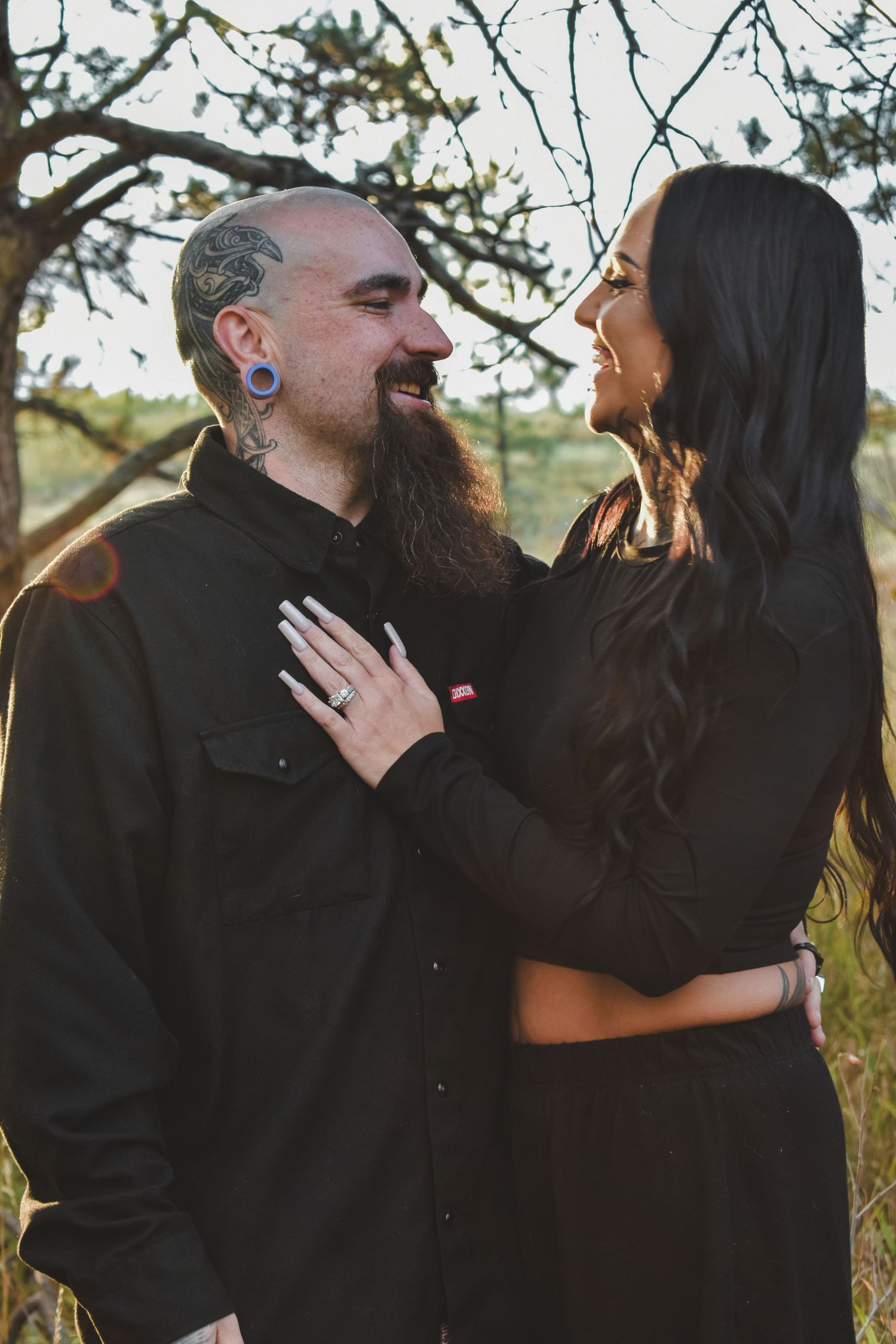 Bearded tattooed man with ear gauges smiling at dark-haired woman during golden hour outdoor couples session.