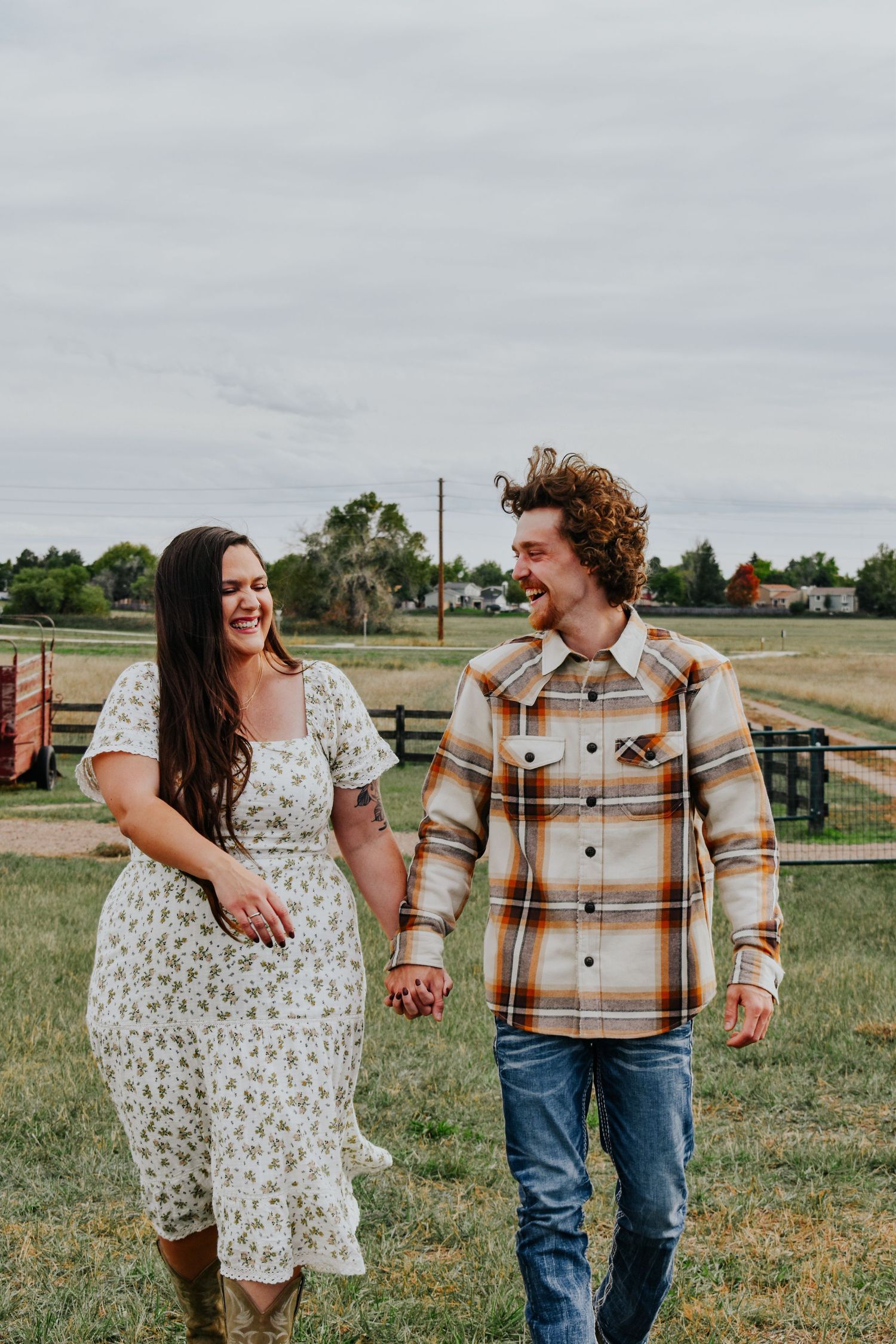 Smiling couple holds hands walking through a farm field, woman in floral dress and man in plaid shirt.