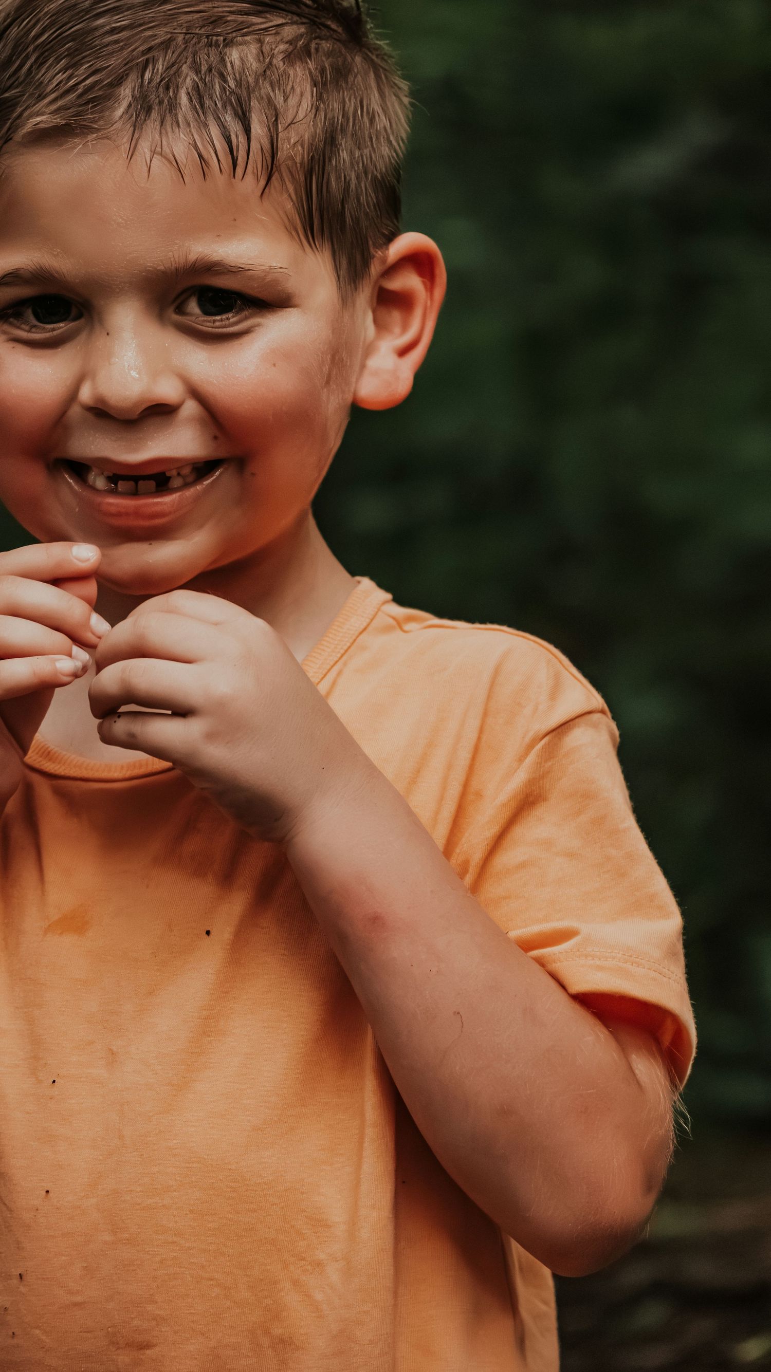 Happy young boy in an orange t-shirt smiles while holding something small in his hands outdoors.