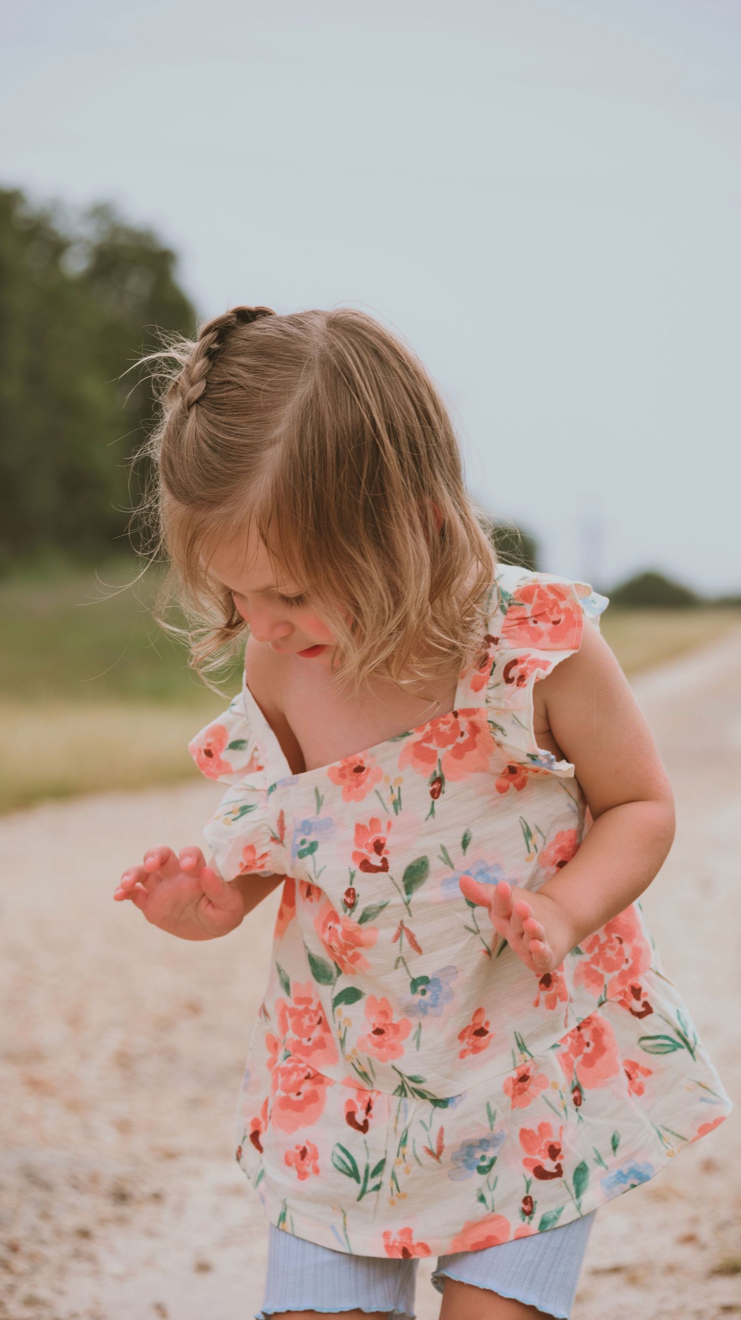Sweet little girl in floral dress looks down while standing on a sunny dirt path.