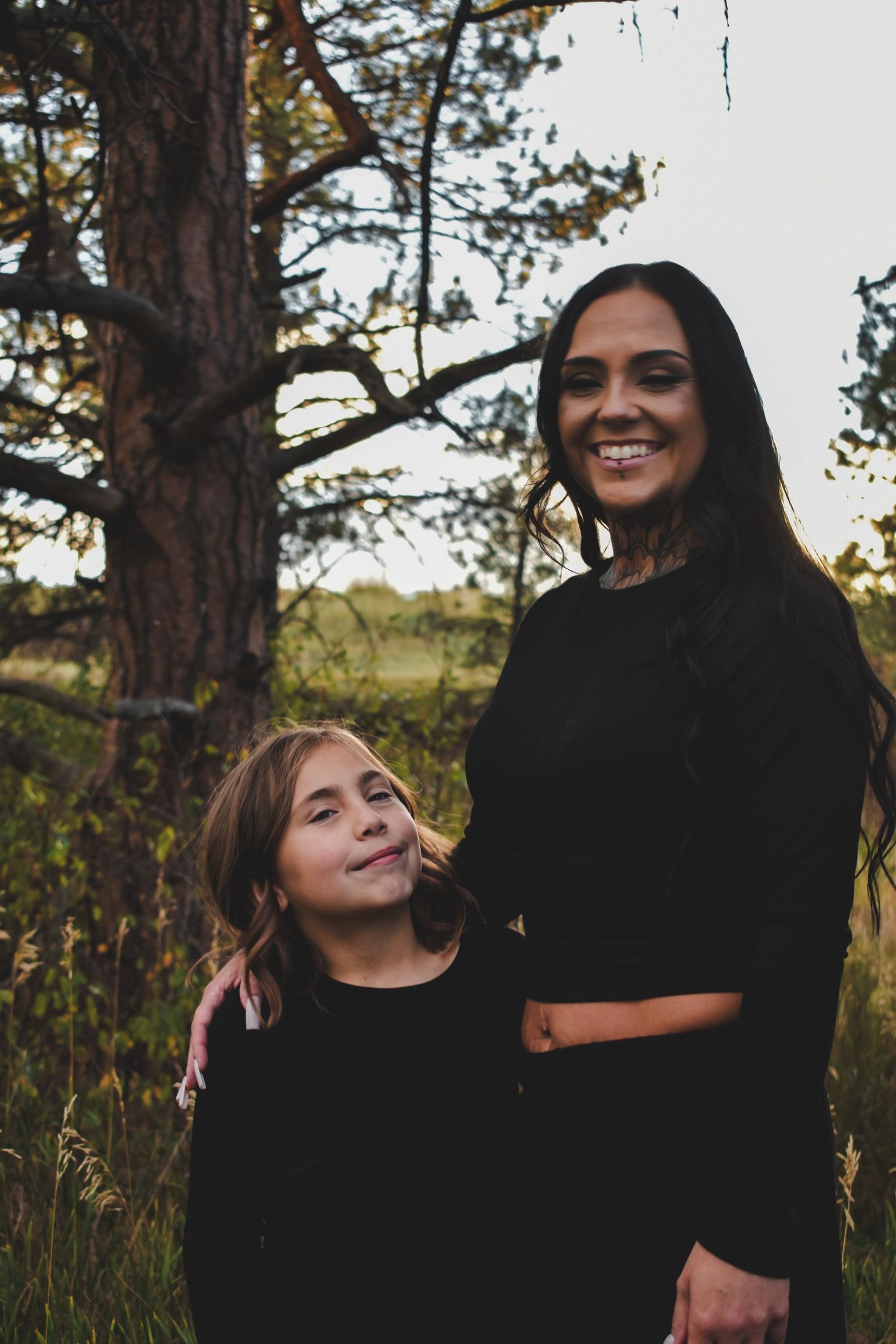 Smiling mother and daughter in black outfits posing together near a tree at sunset.