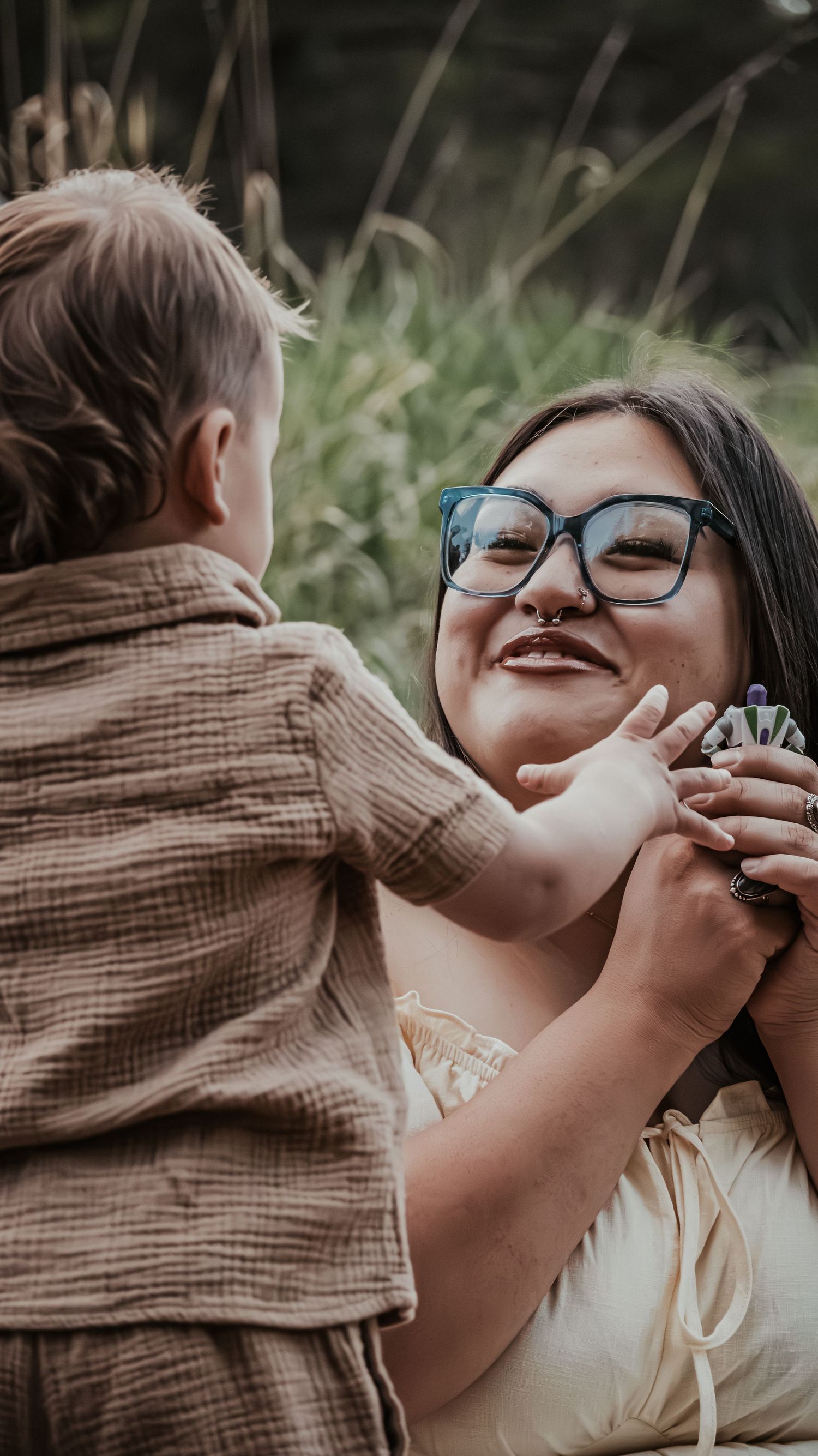 Smiling woman with glasses joyfully interacting with young toddler boy outdoors in natural setting.