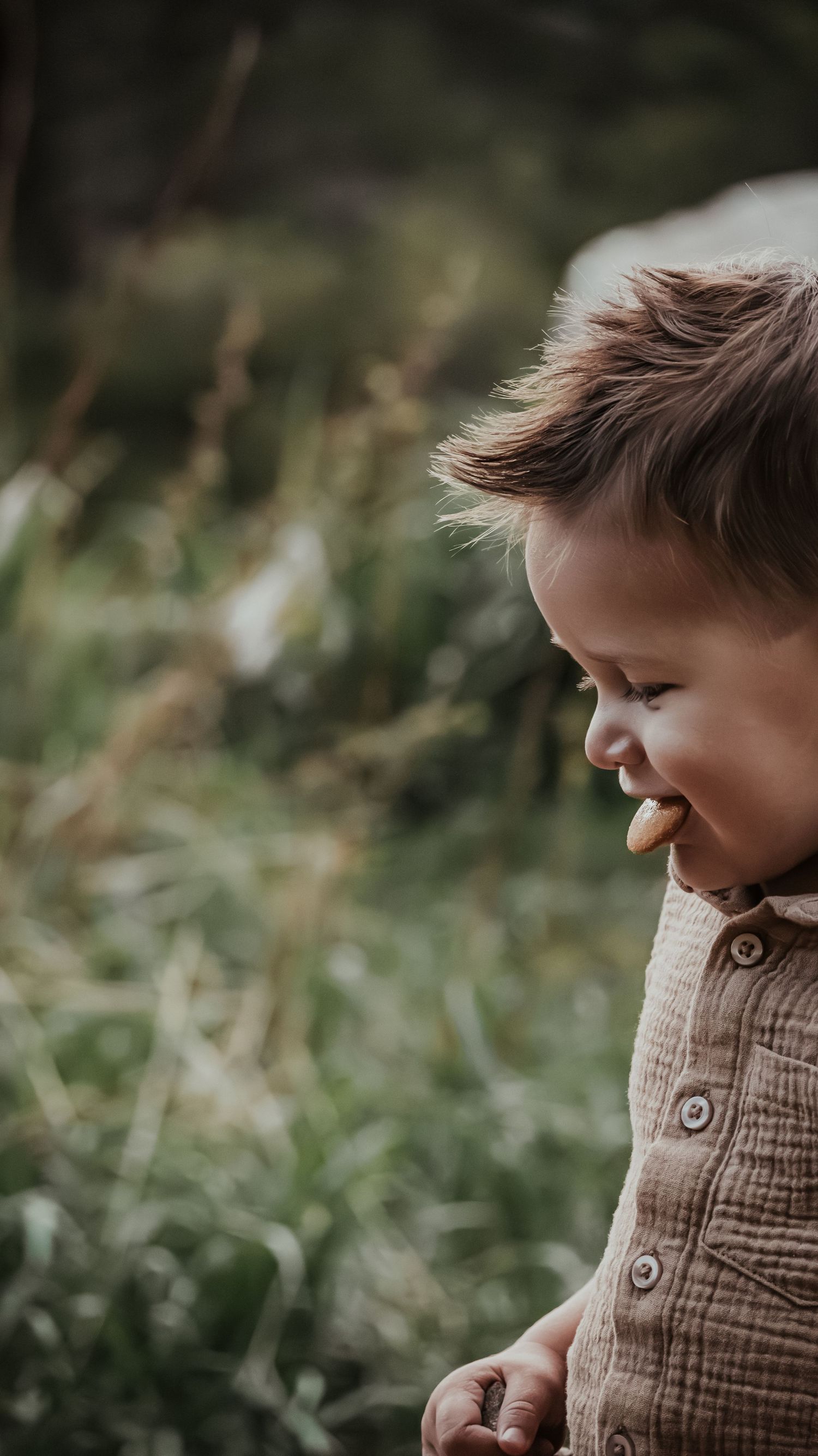 Smiling toddler boy with windswept hair wearing a brown linen shirt laughing outdoors in a field.