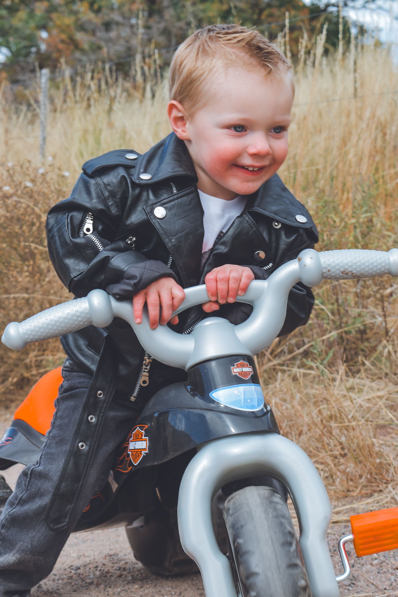 Happy toddler in Harley-Davidson leather jacket gripping handlebars of toy motorcycle outdoors.