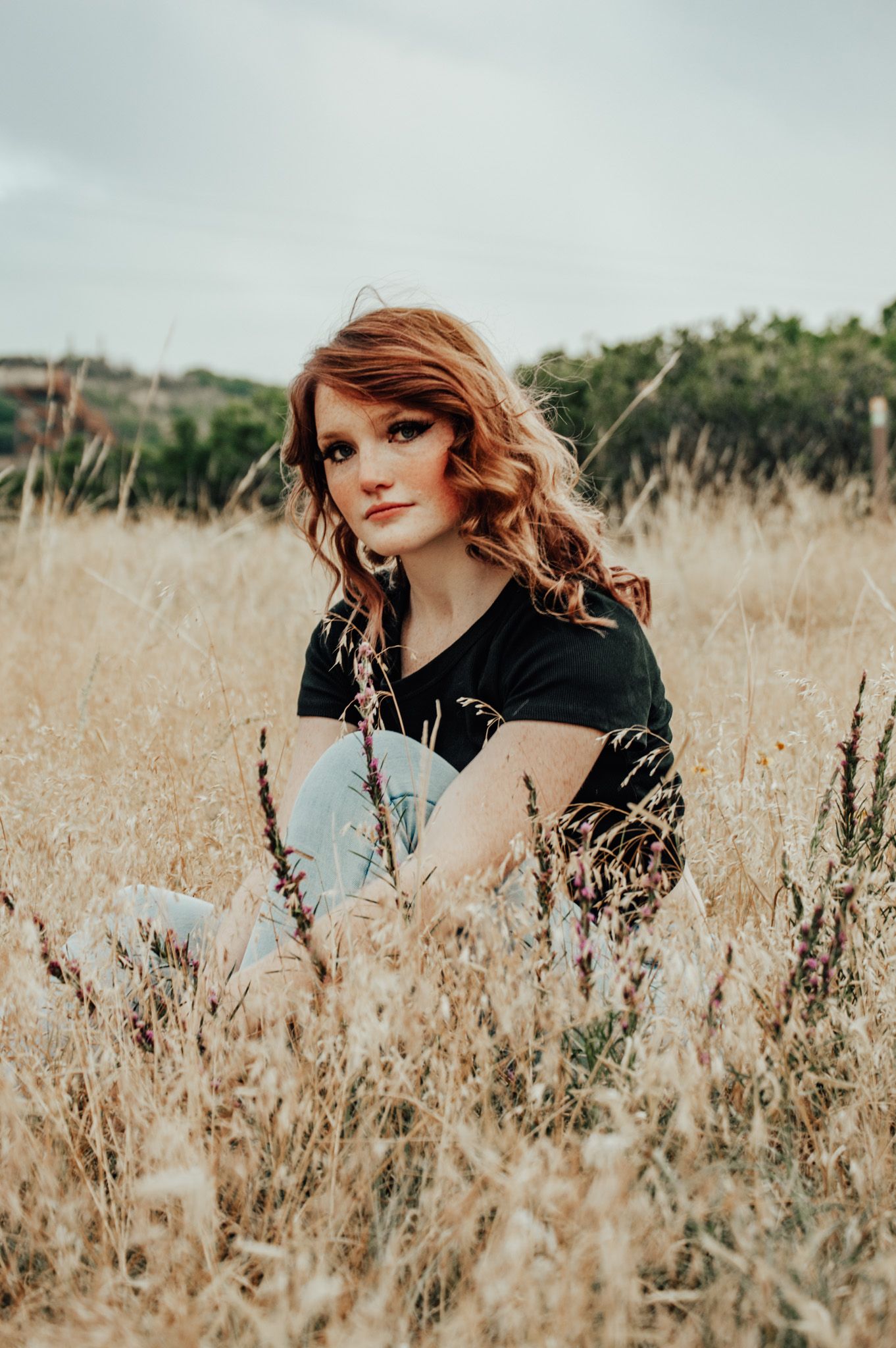 Redhead woman sitting in golden dry grass field wearing black t-shirt and light blue jeans outdoors for senior photos.
