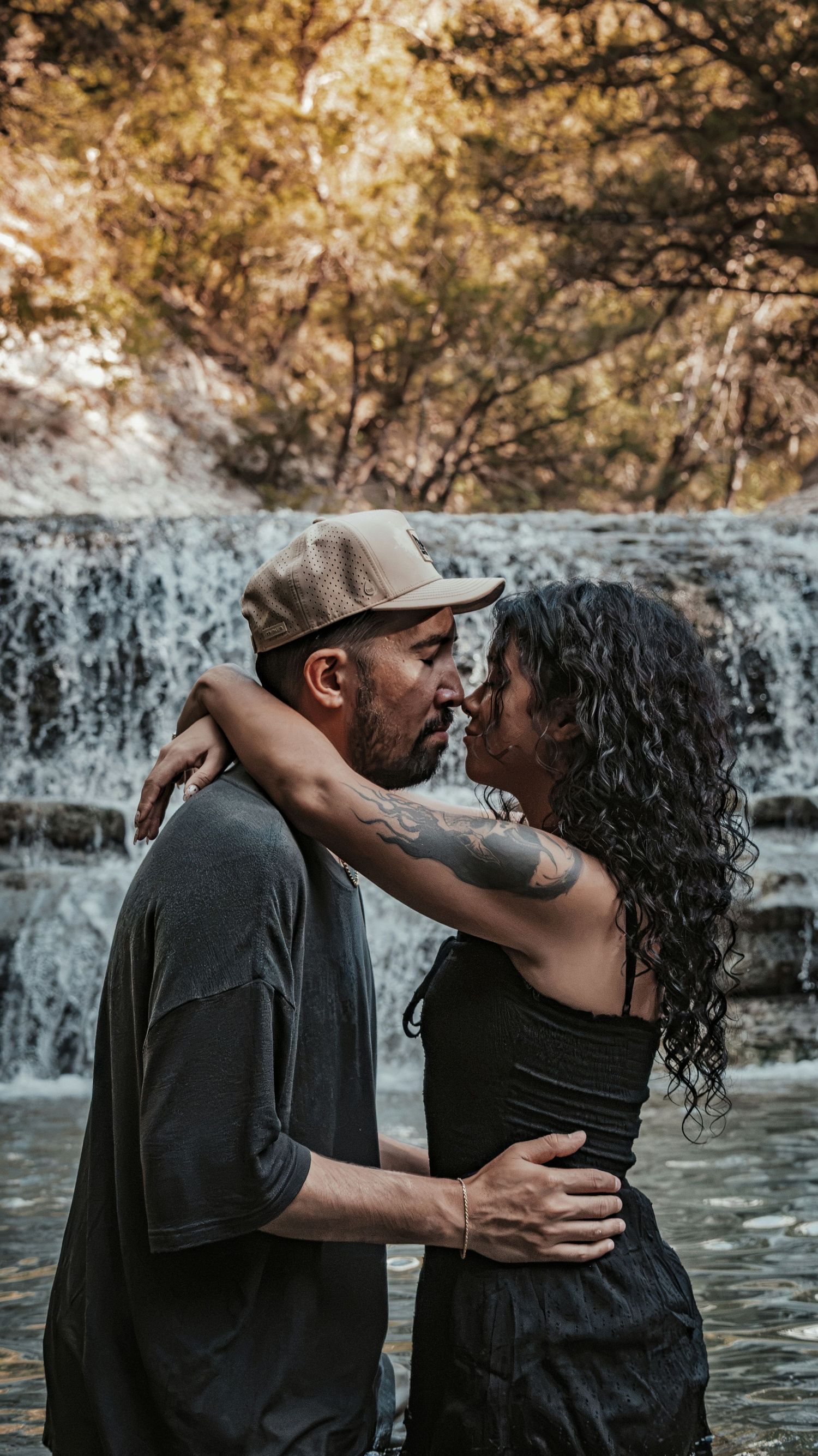 Tattooed couple sharing a romantic kiss in front of a waterfall.