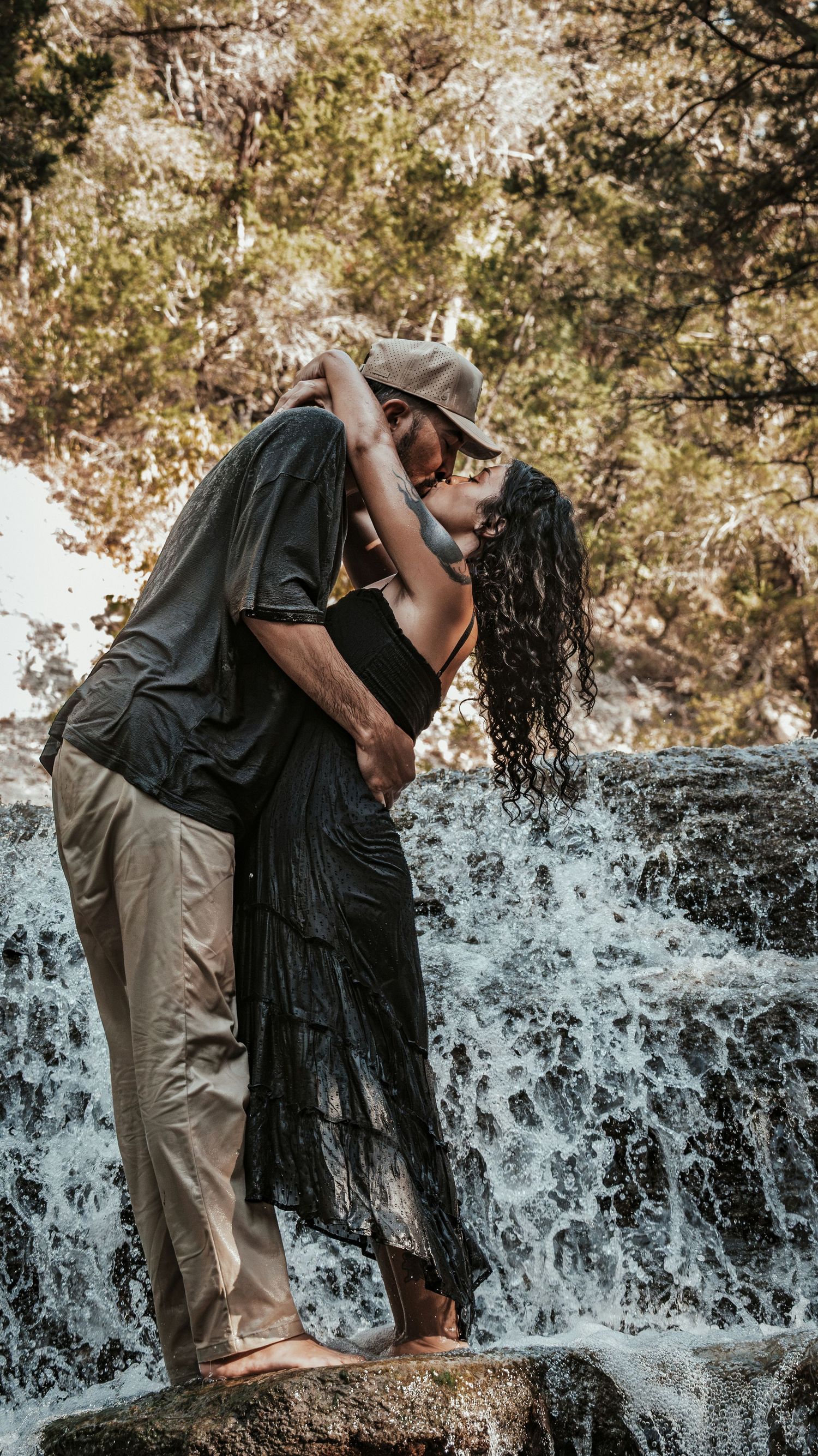 Couple sharing a romantic kiss while standing barefoot on rocks before a waterfall.