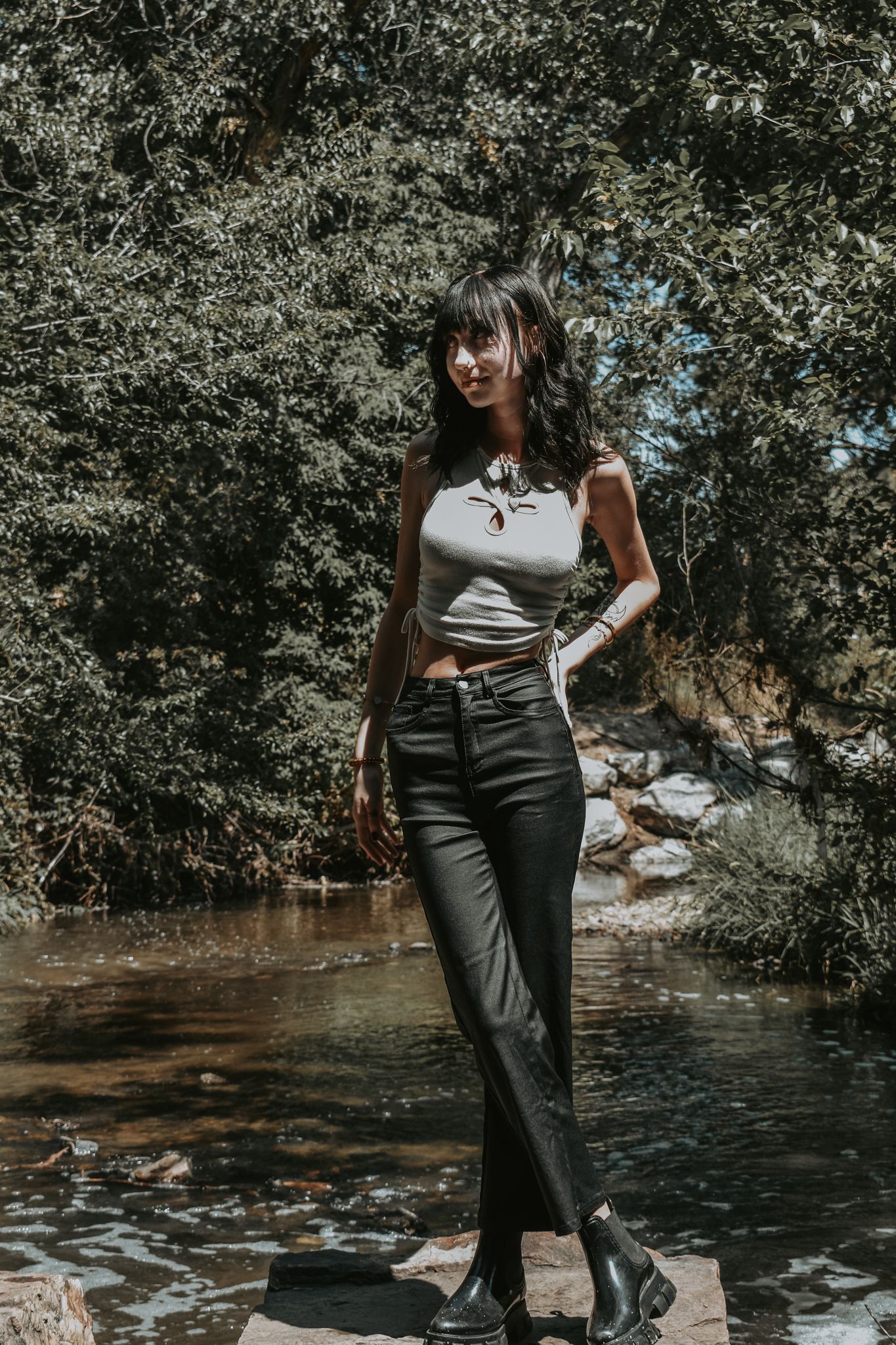 Stylish young woman in white crop top and black jeans stands near a rocky creek surrounded by trees for senior photos.