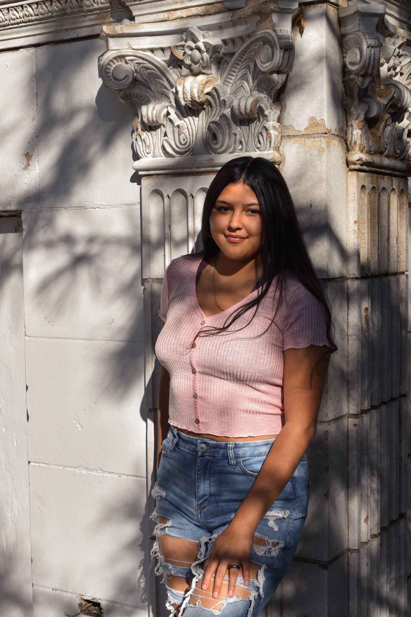 Young woman in pink crop top and ripped jeans poses by ornate white stone wall for senior photos.