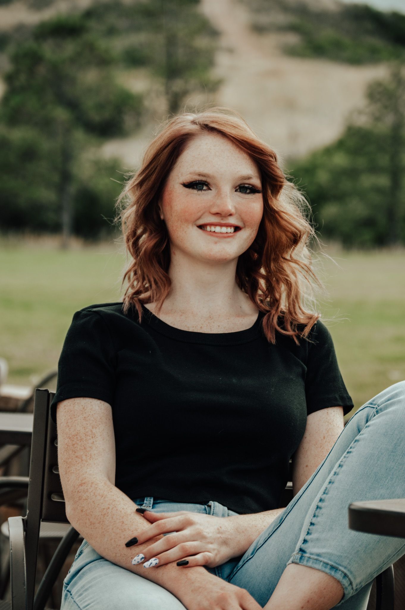Smiling redhead woman with freckles in black tee and light jeans sitting outdoors with green hills behind for senior photos.