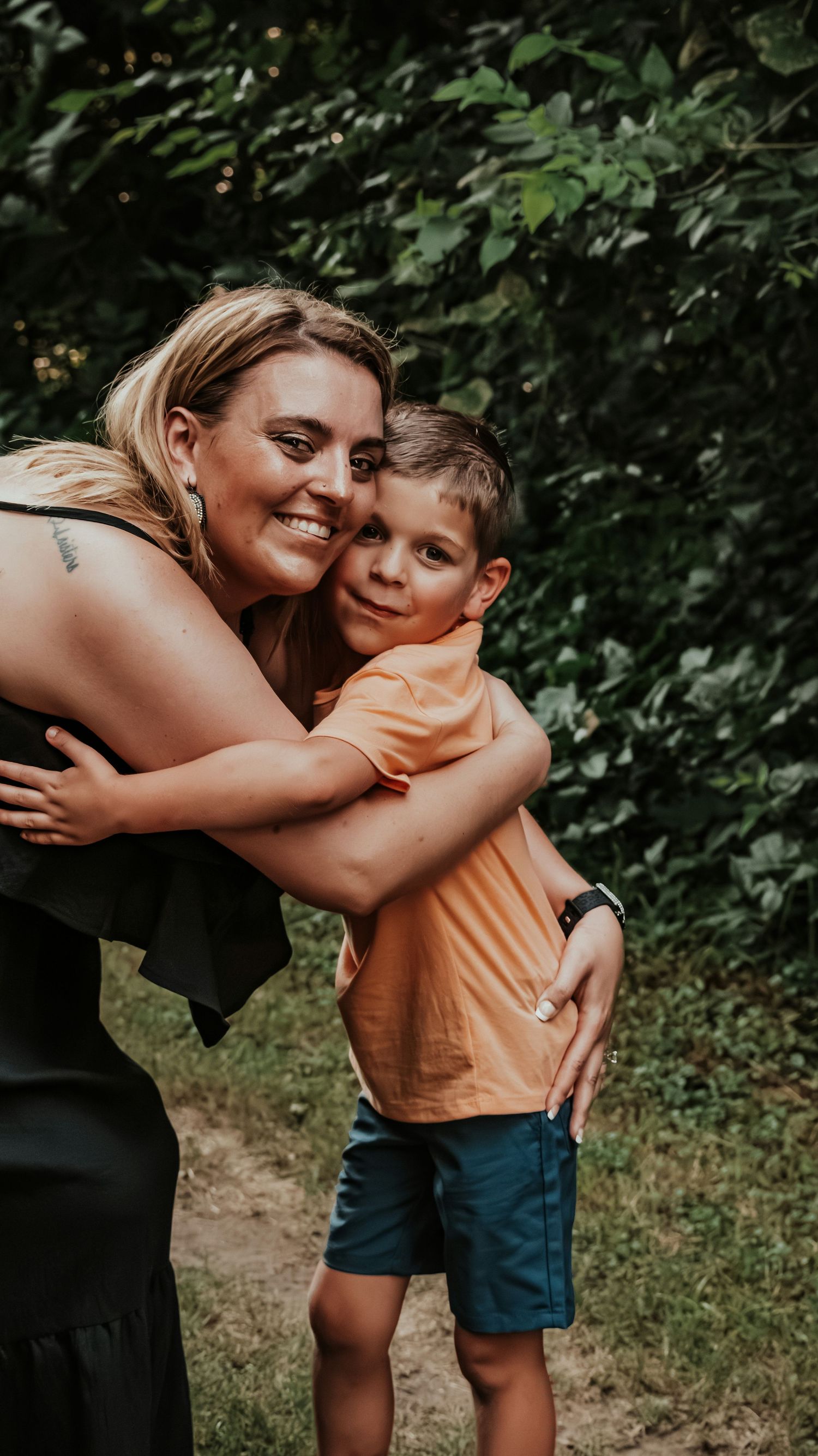 A smiling woman hugs a young boy in an orange shirt outdoors, surrounded by lush green trees.