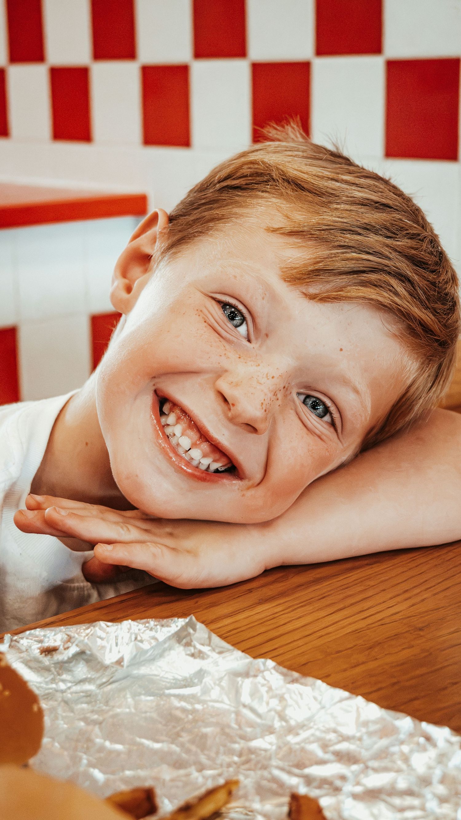 Smiling freckled red-haired boy resting chin on hands at a wooden table. Family Photographer in Pflugerville, Tx