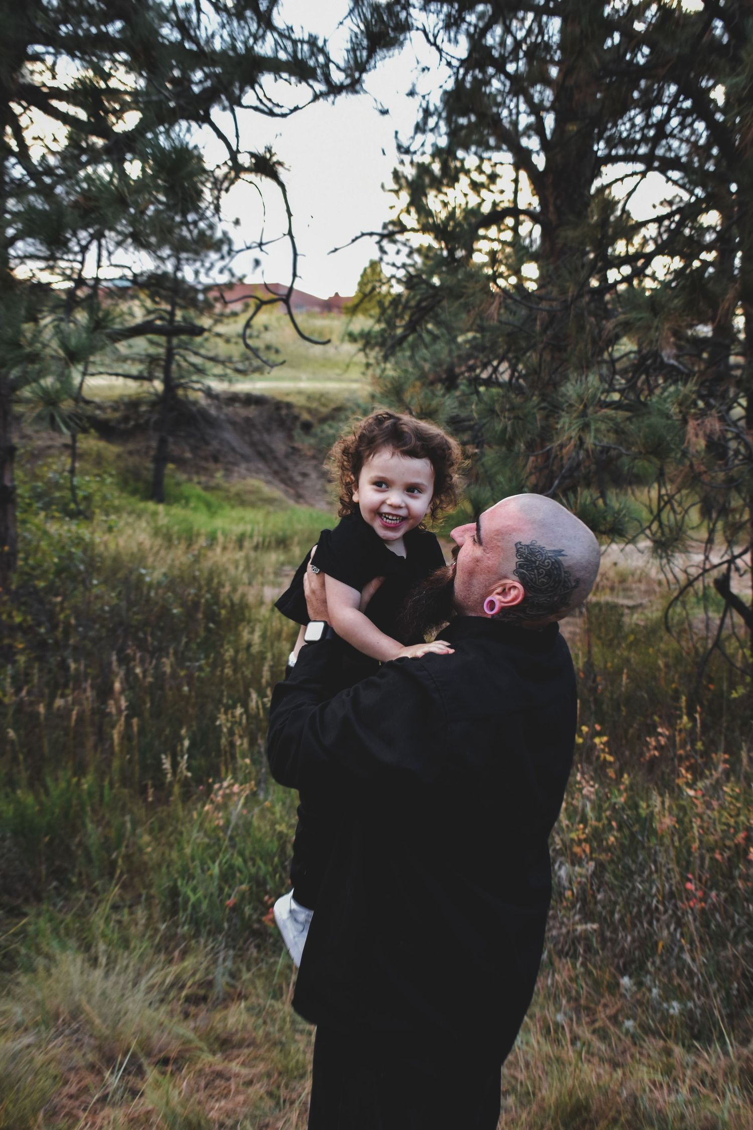 A bald bearded man lovingly holds a curly-haired toddler in black outdoors among pine trees. Family Photographer in Pflugerville, Tx