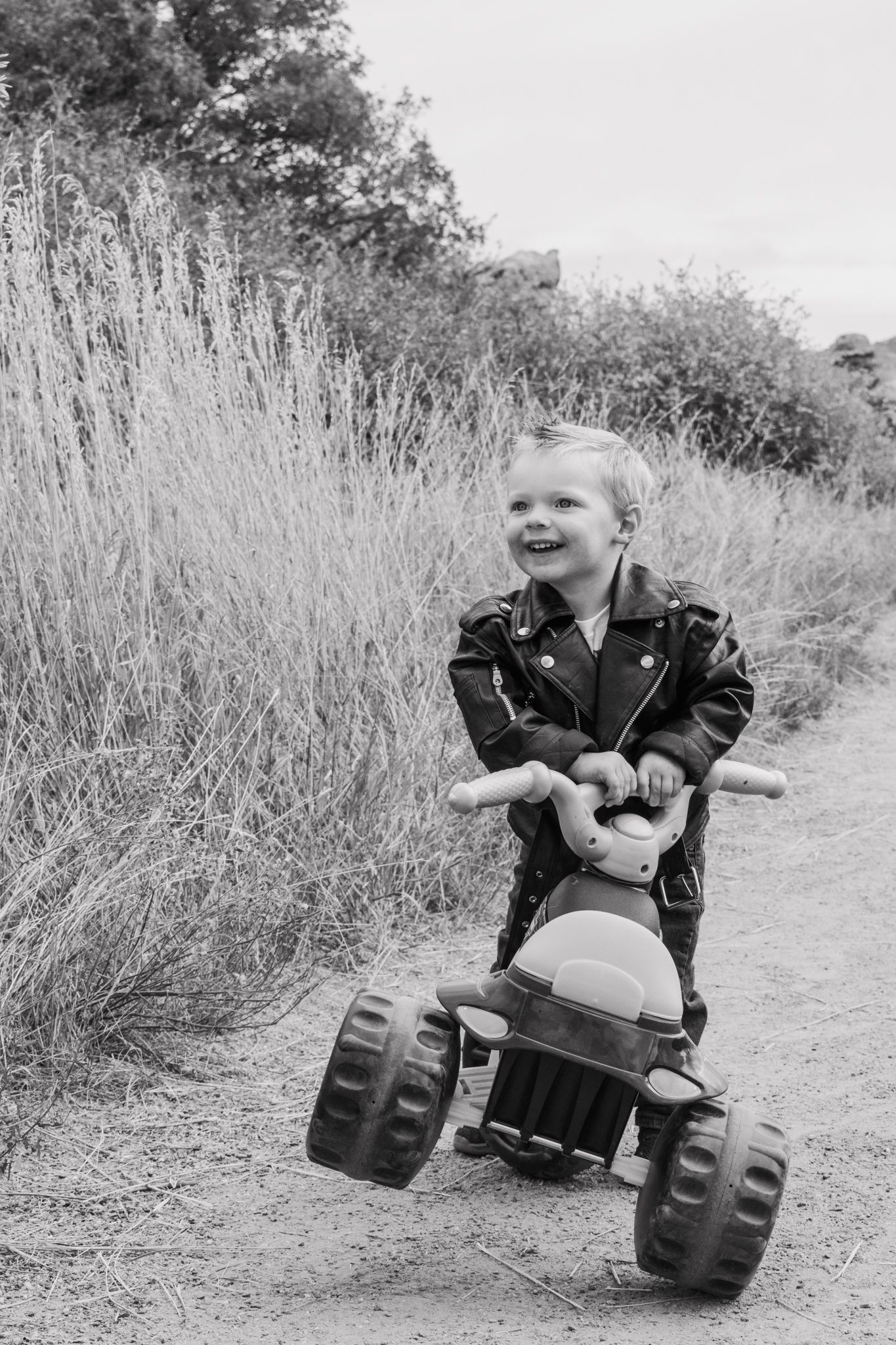 Happy toddler in leather jacket rides toy tricycle on dirt path surrounded by tall grass. Family Photographer in Pflugerville, Tx