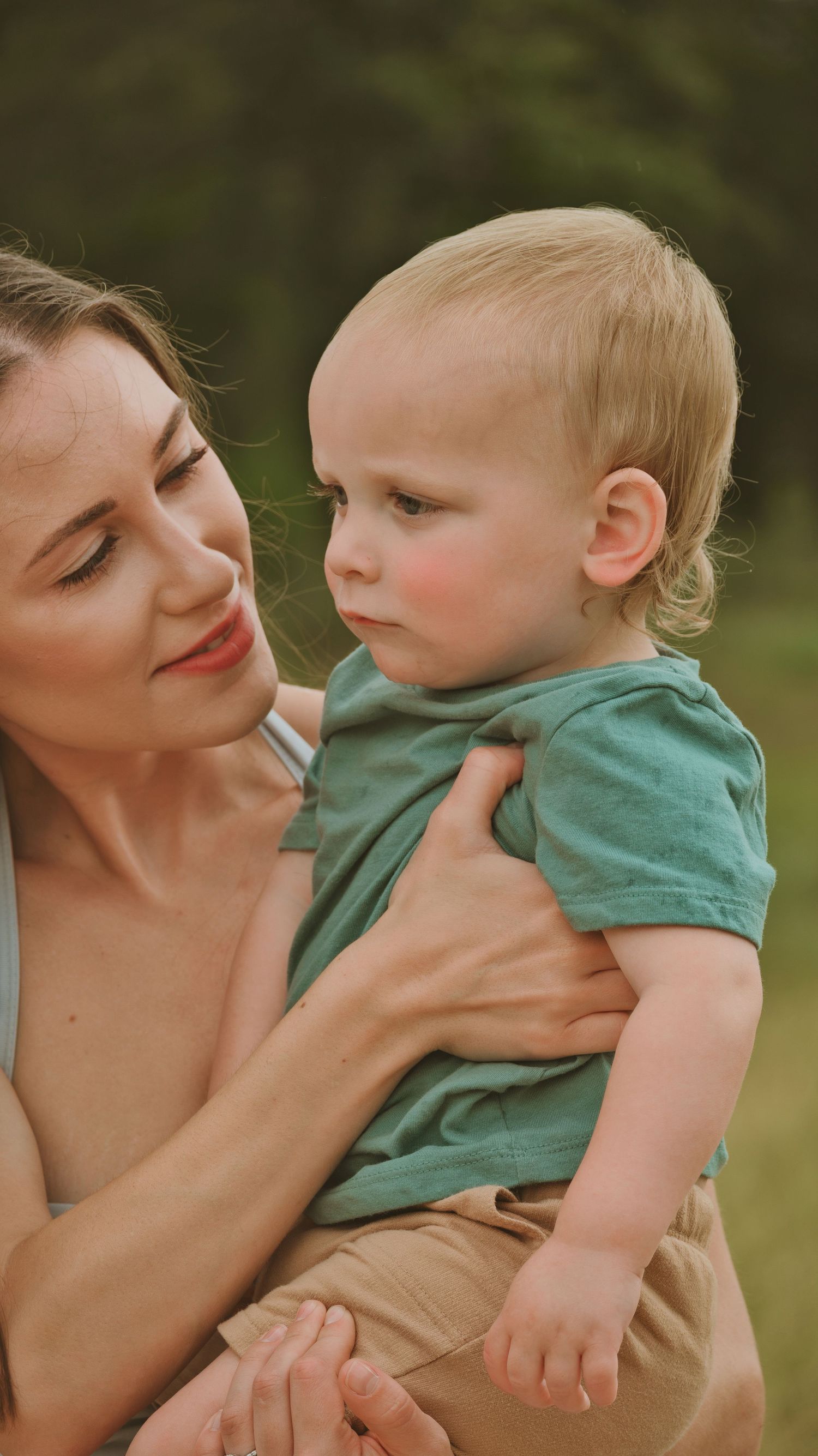 A mother tenderly holds her blonde toddler boy in a green tee outdoors.