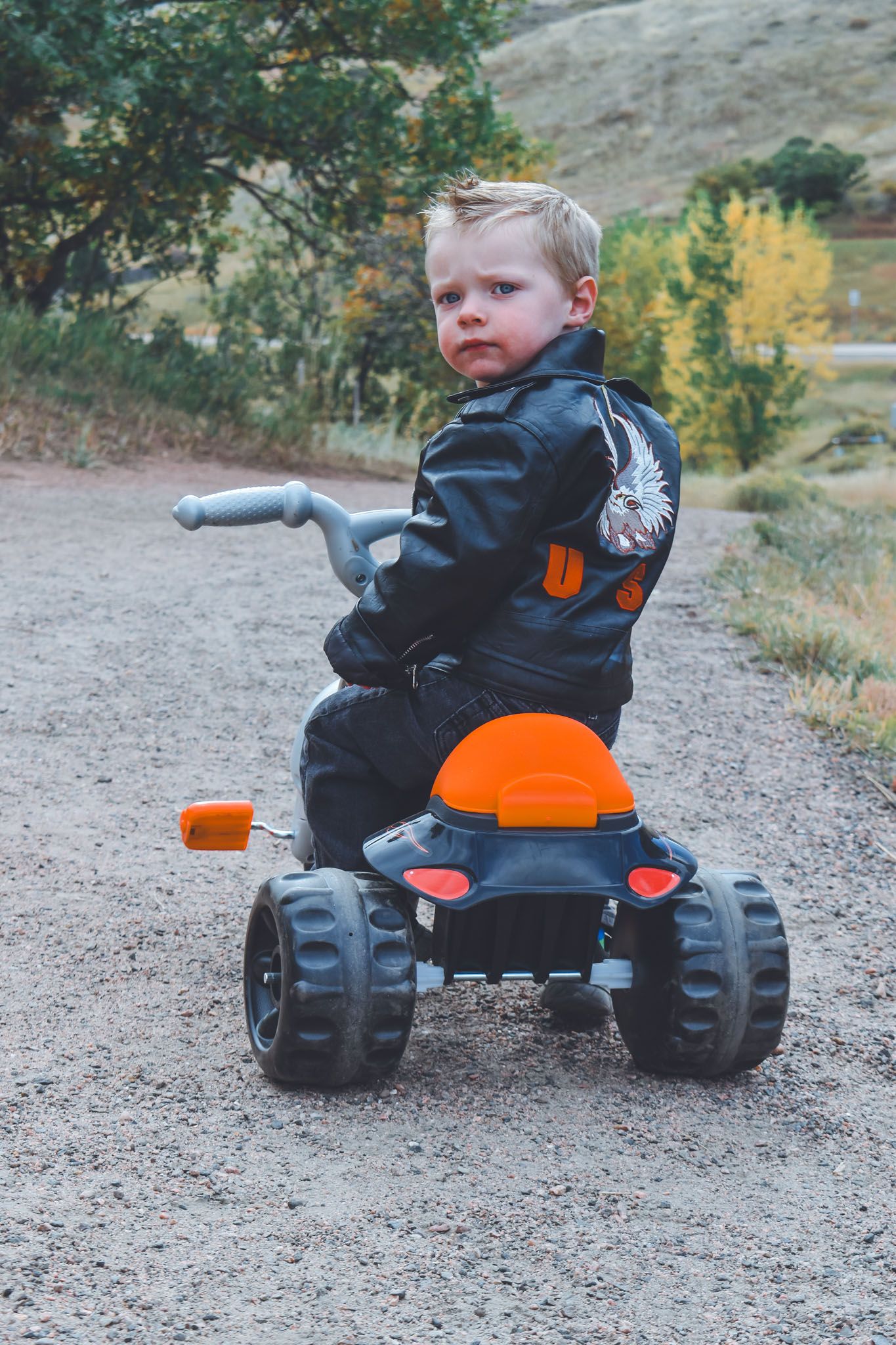 Toddler boy in a black leather jacket sitting on a toy ATV on a gravel path.