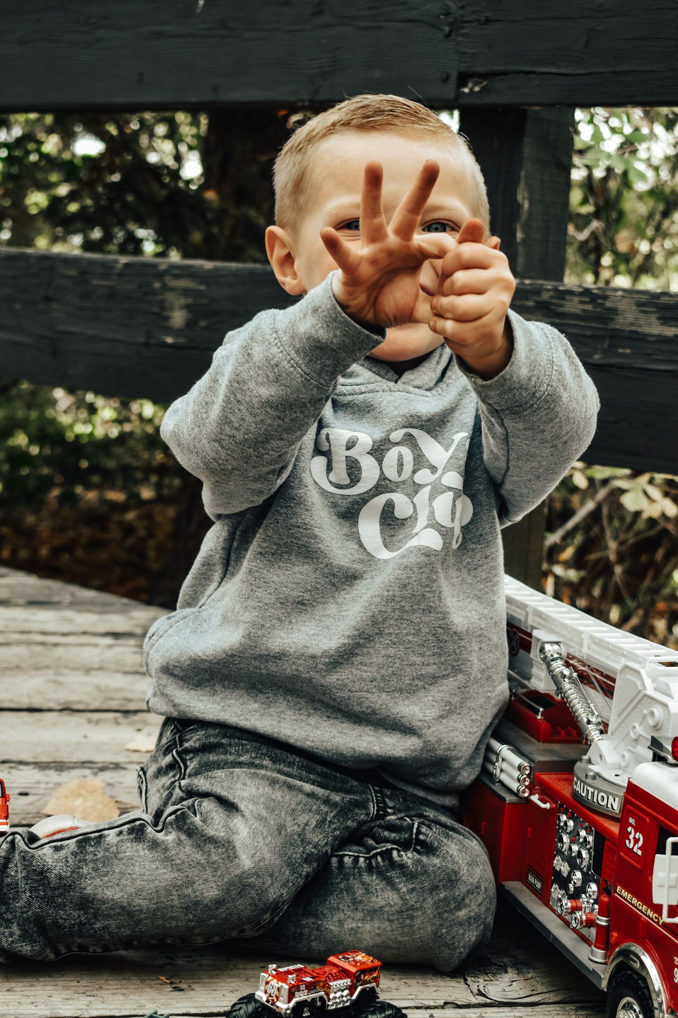 Toddler boy in grey Boys Club sweatshirt holding up fingers while sitting with toy fire trucks on wooden deck.