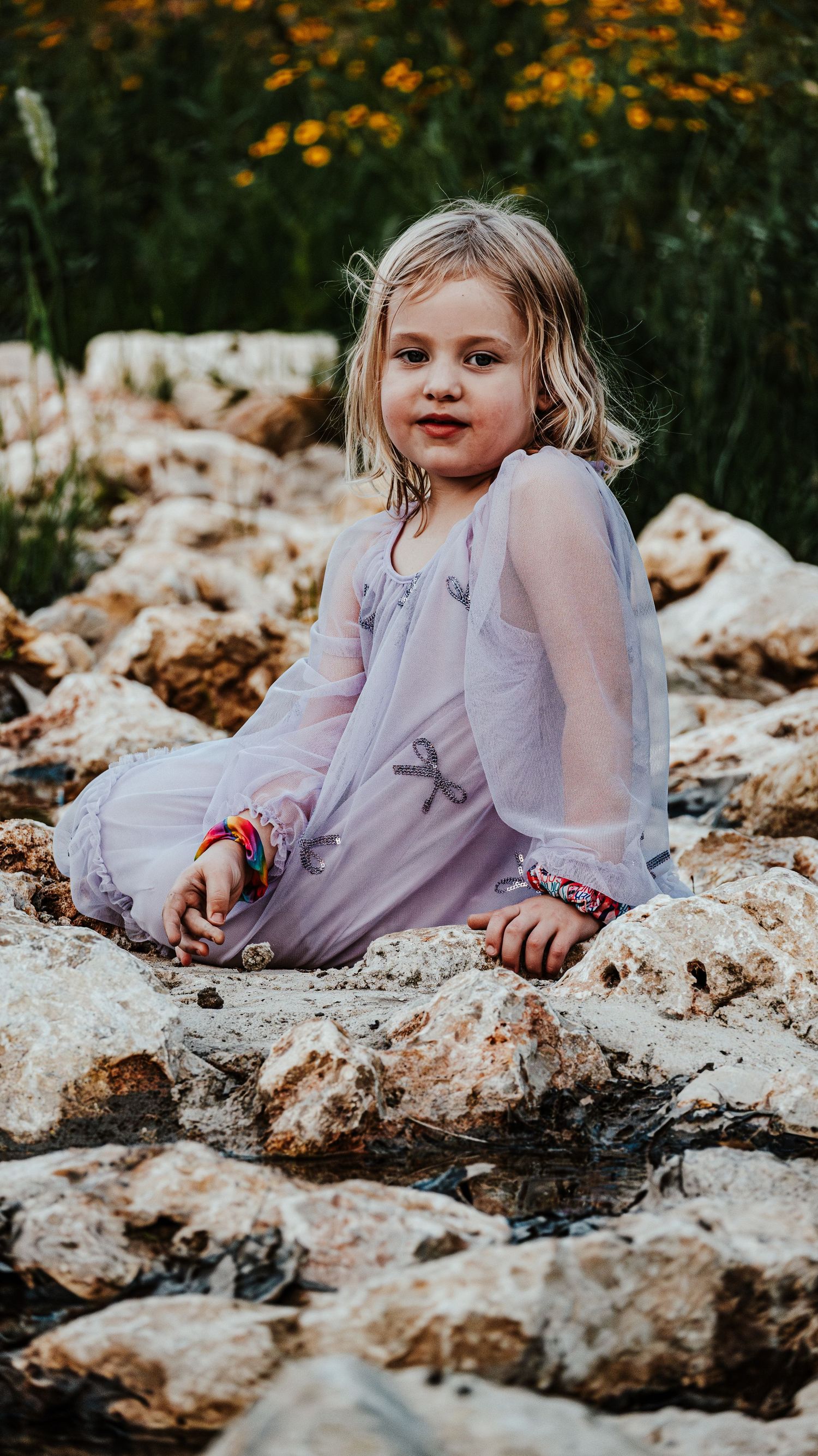 Young girl in purple dress sitting on rocks near water with yellow flowers behind her.