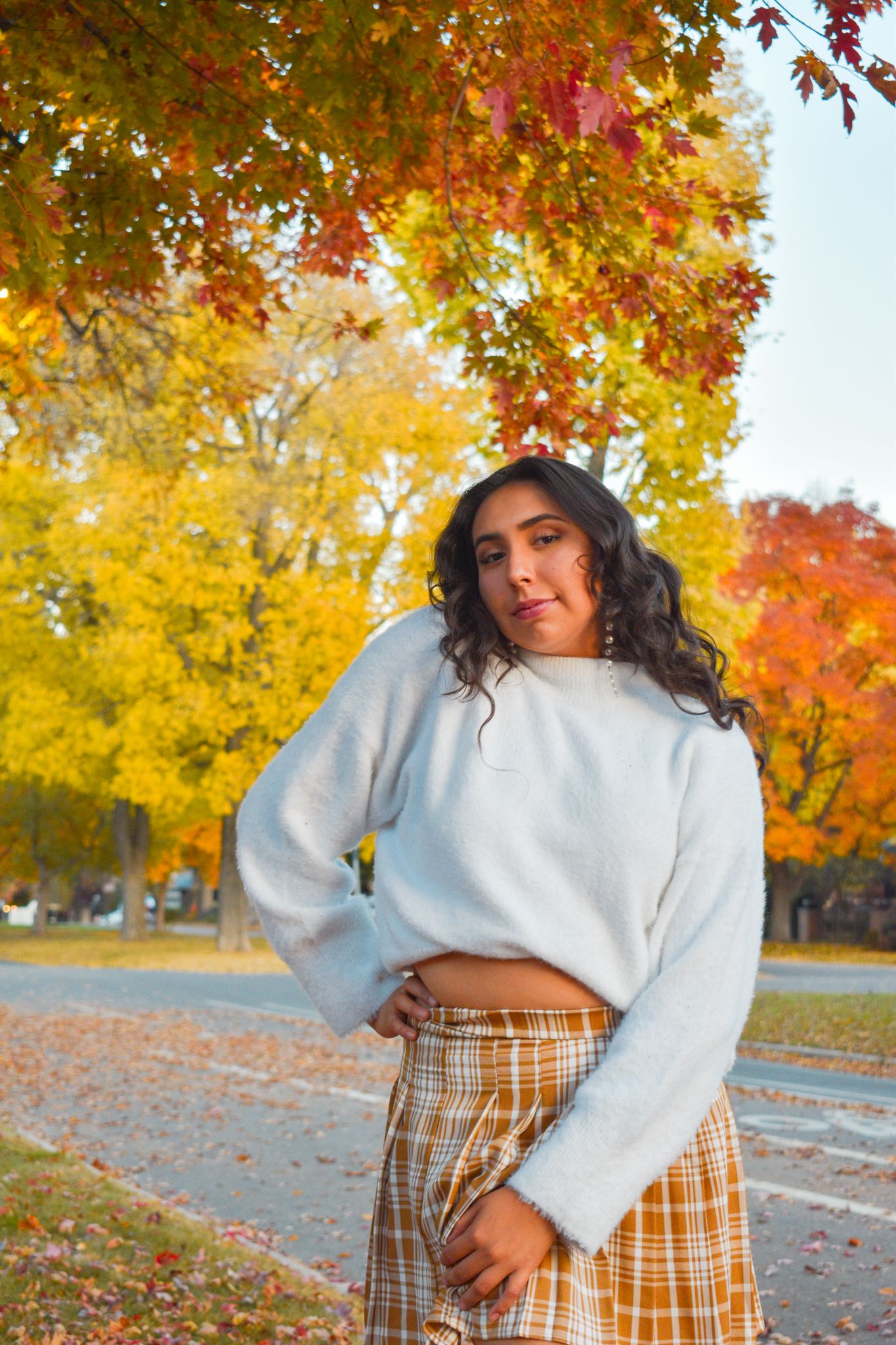 Young woman in white crop sweater and plaid skirt posing under vibrant yellow and red autumn trees for senior photos.