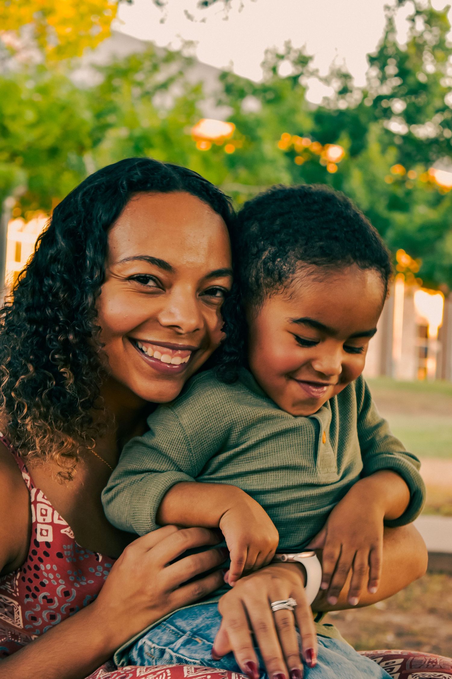 Smiling African American mother hugs her young son affectionately while sitting outdoors in a green park at sunset.